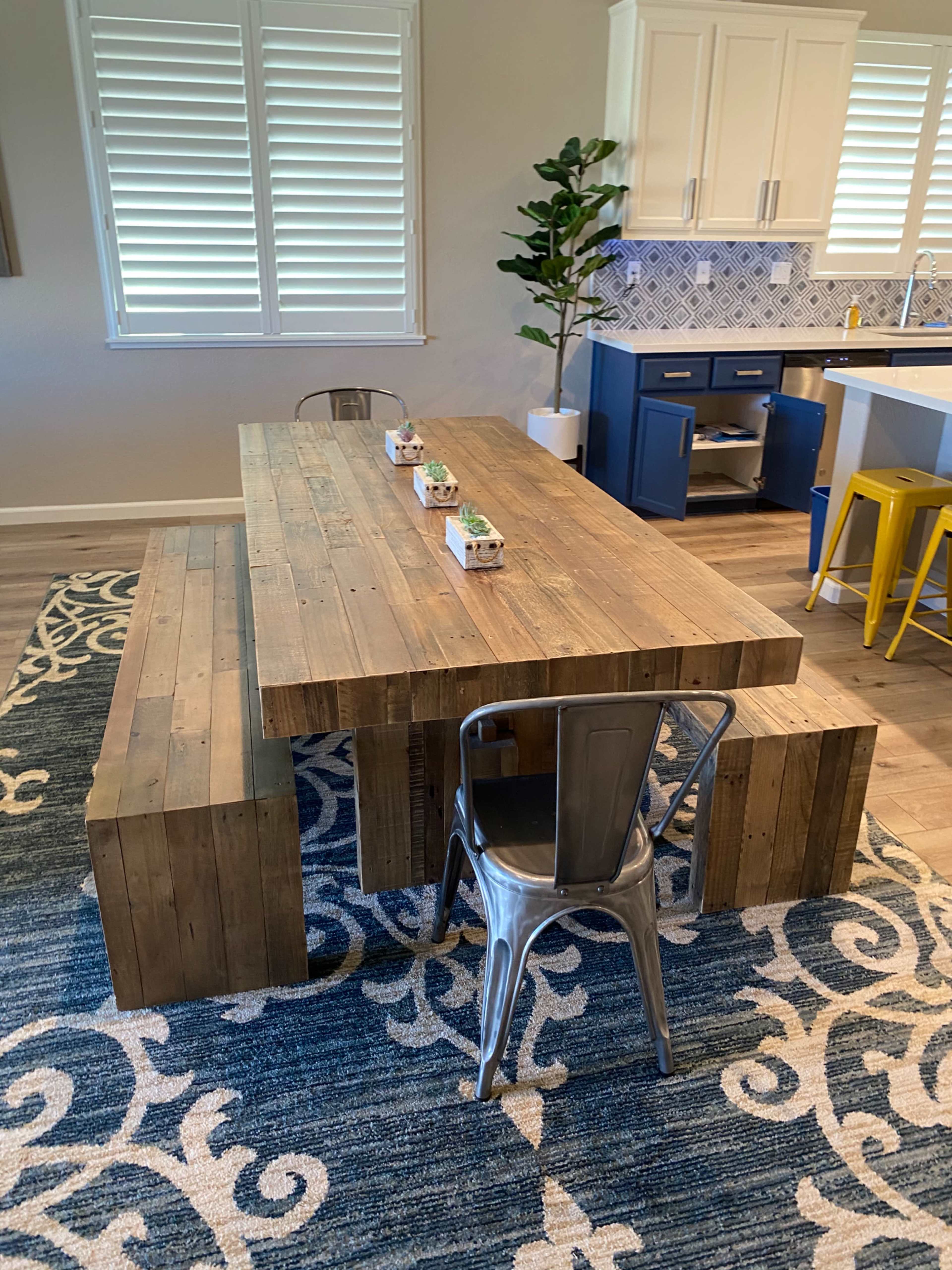 A wooden dining table with metal chairs is placed on a patterned rug in a modern kitchen setting.