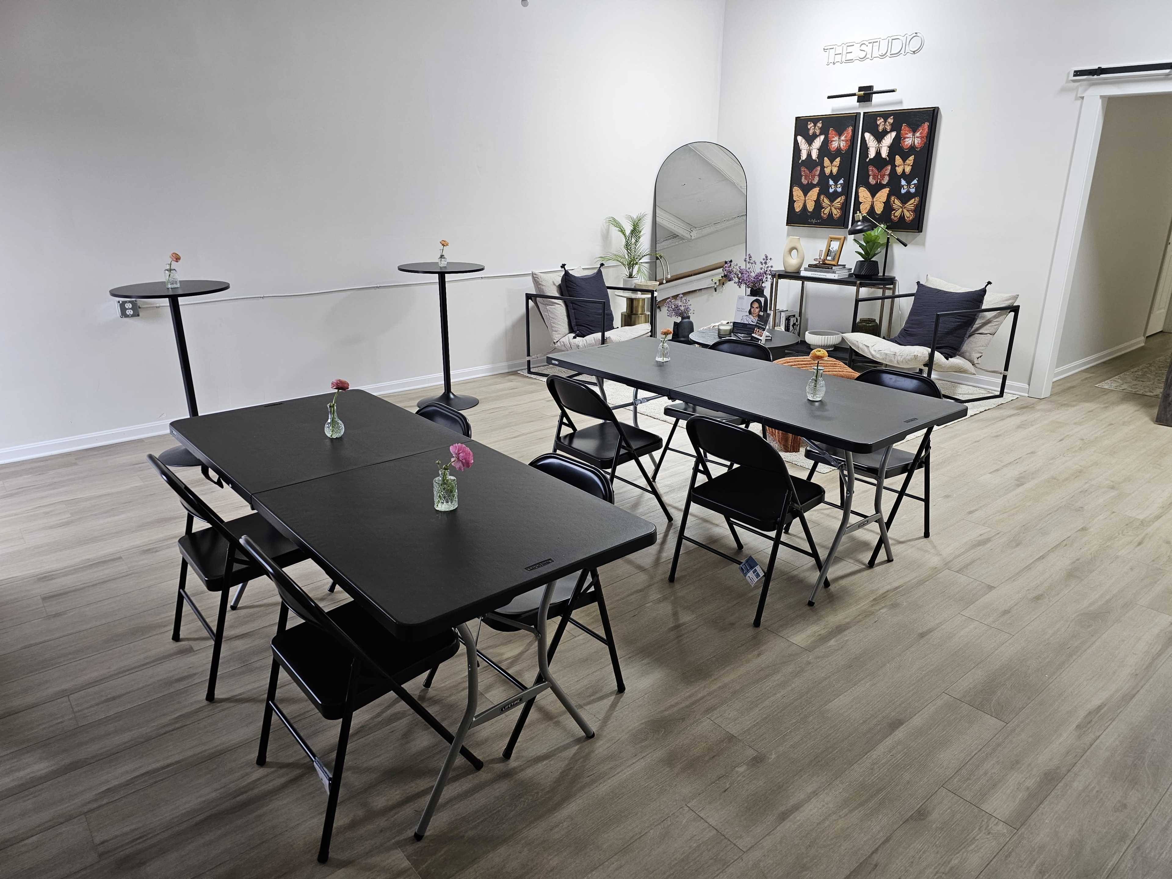 The image shows a light-colored room with several black folding tables arranged in a grid, each adorned with small vases containing flowers.