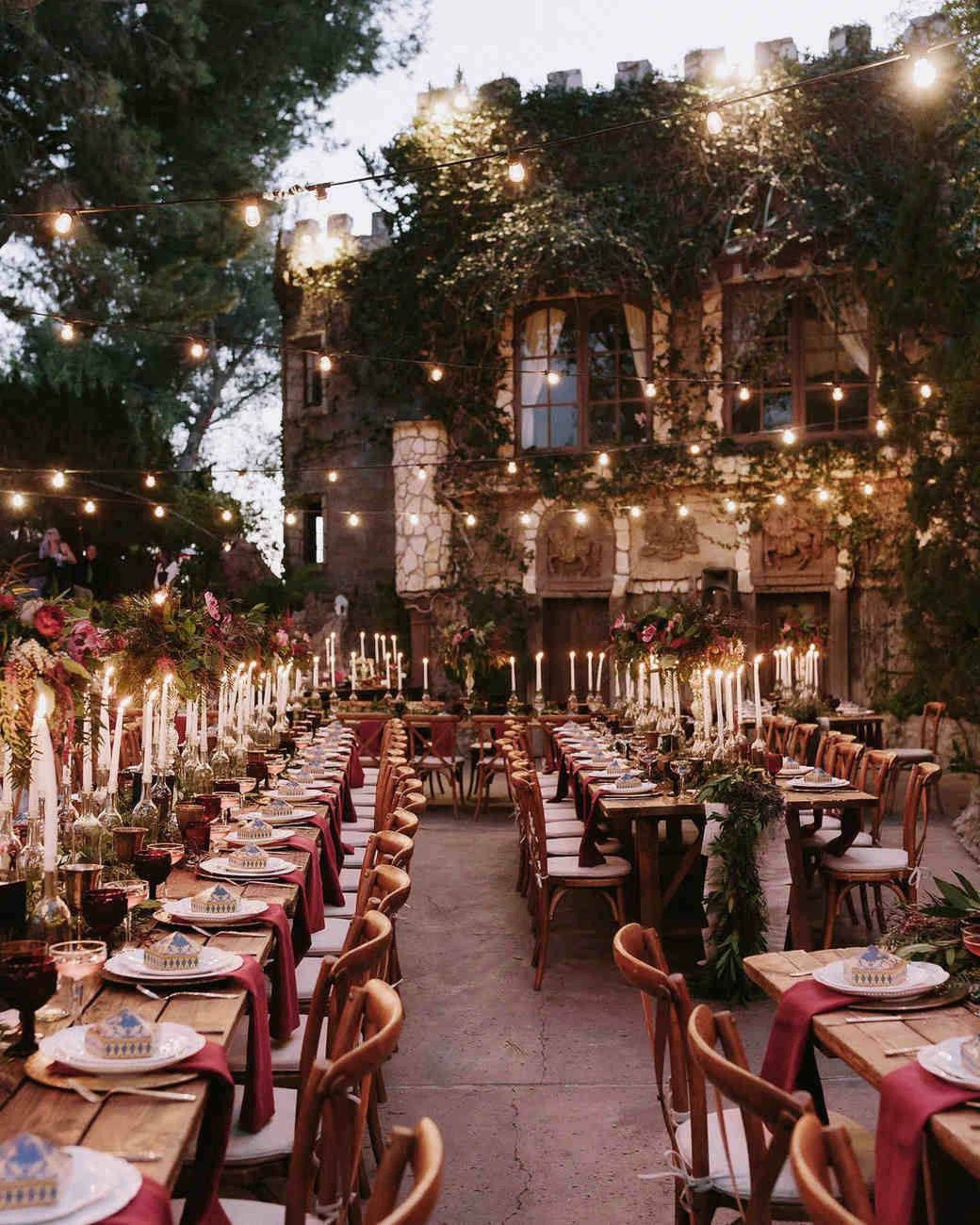 An outdoor dinner setting features long tables adorned with candles and floral centerpieces, illuminated by string lights against the backdrop of a castle-like structure.
