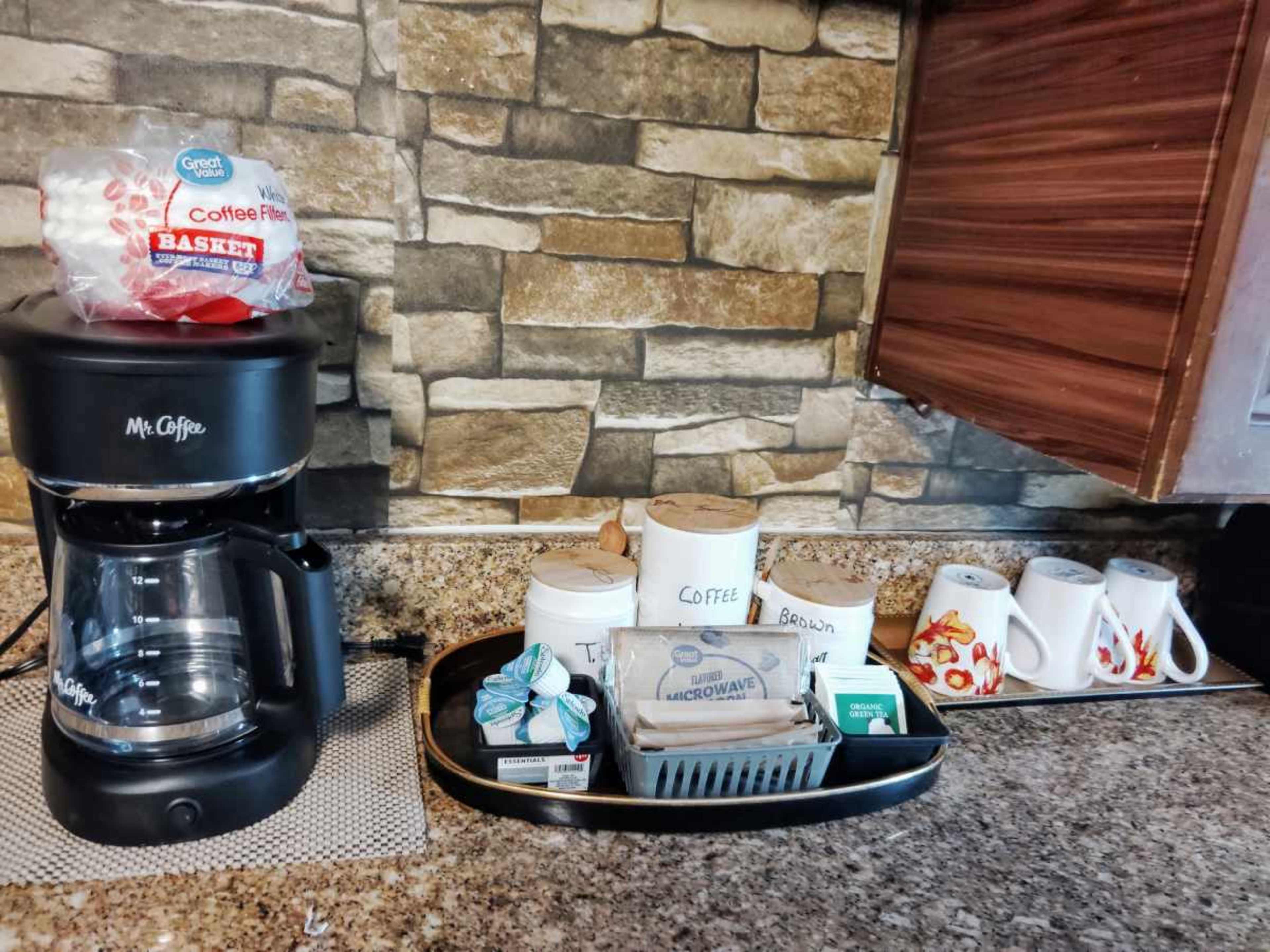 The image shows a countertop with a coffee maker, a basket of coffee filters, labeled containers for coffee and tea, and several decorative mugs.
