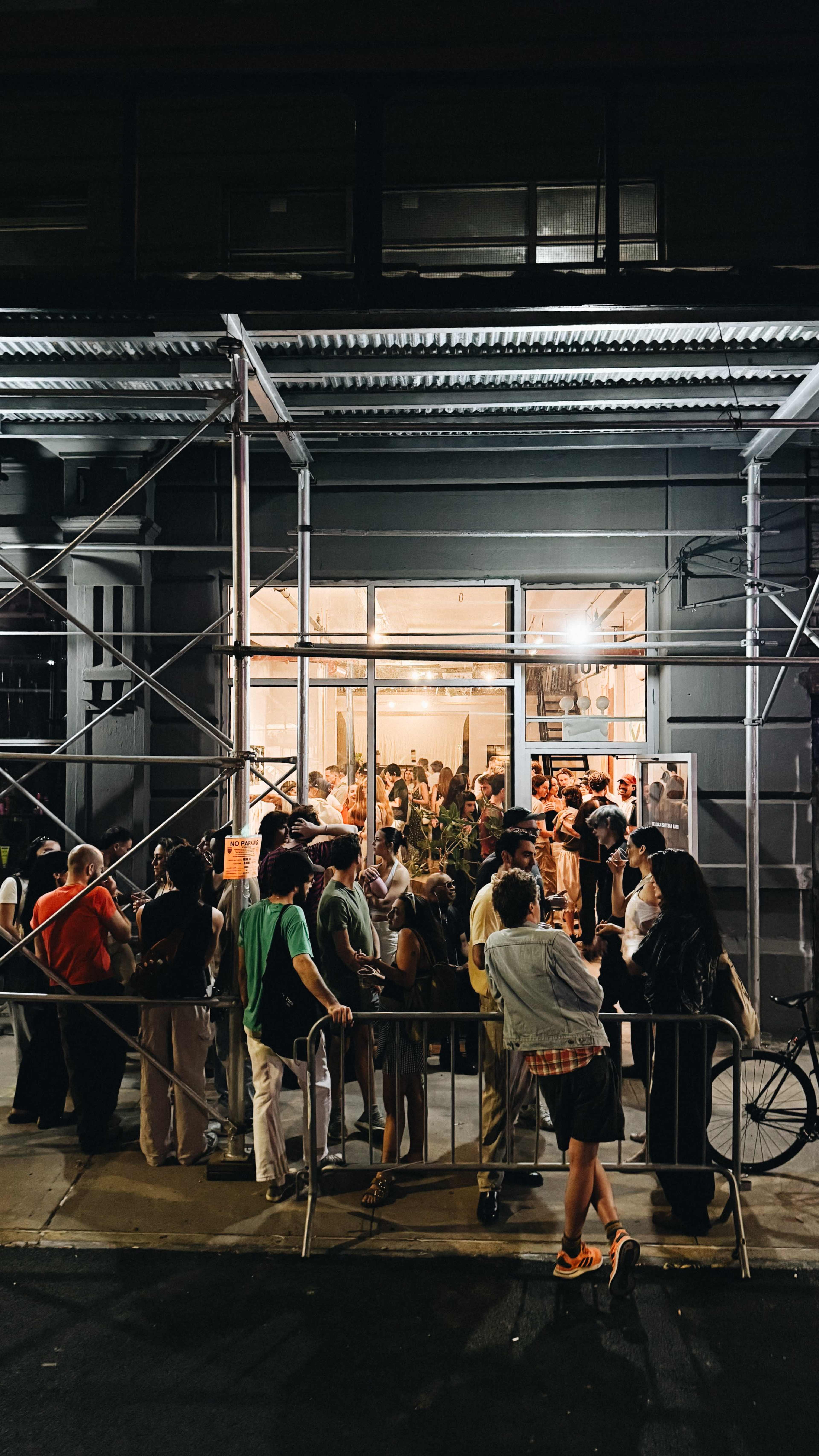 A crowd of people stands outside a building under construction, waiting to enter the well-lit interior.