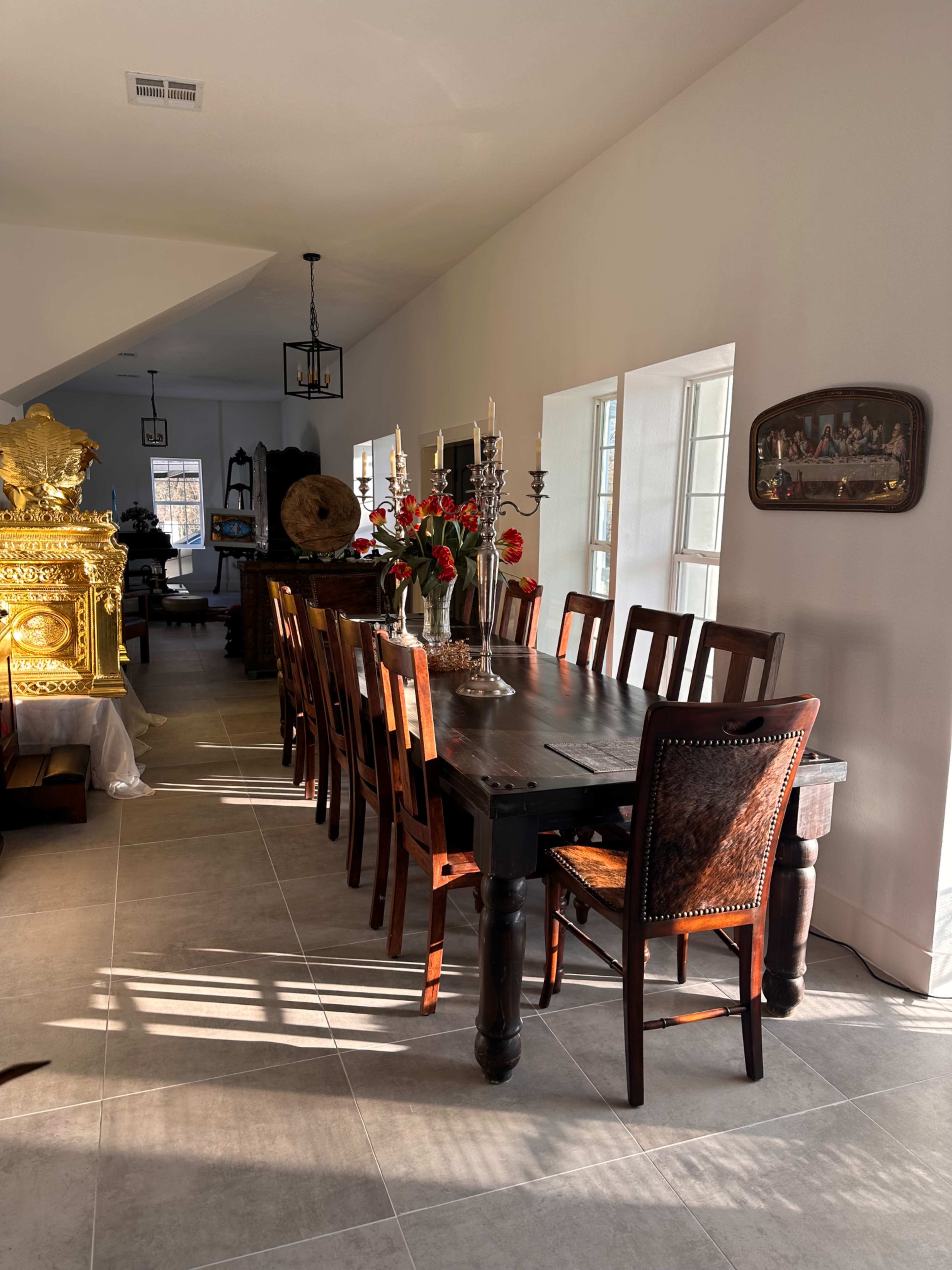A long wooden dining table with multiple chairs is set in a bright room, featuring large windows and decorative elements like flowers and a golden sculpture.