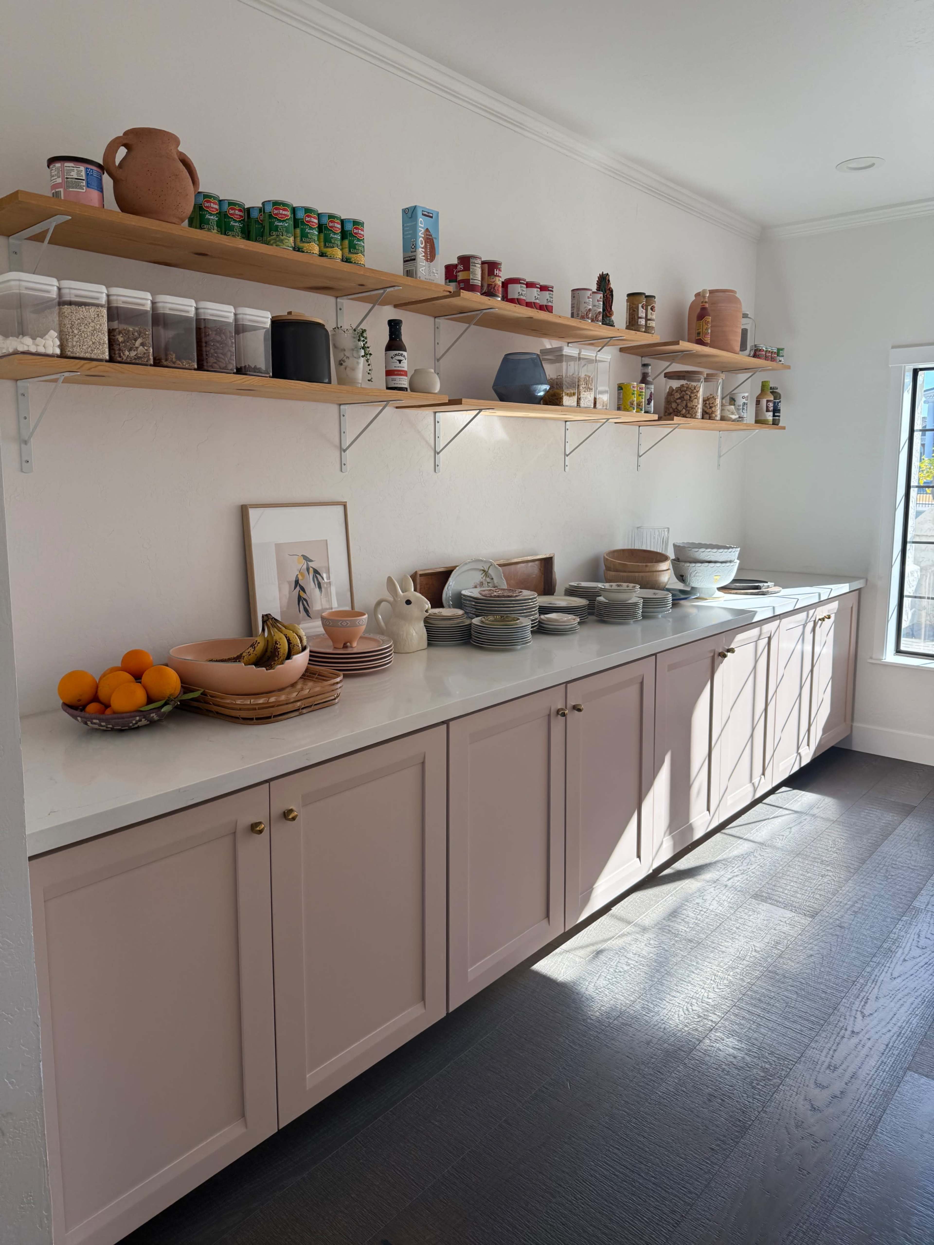 A kitchen countertop features a variety of dishes, fruits, and canned goods neatly arranged on floating shelves above.