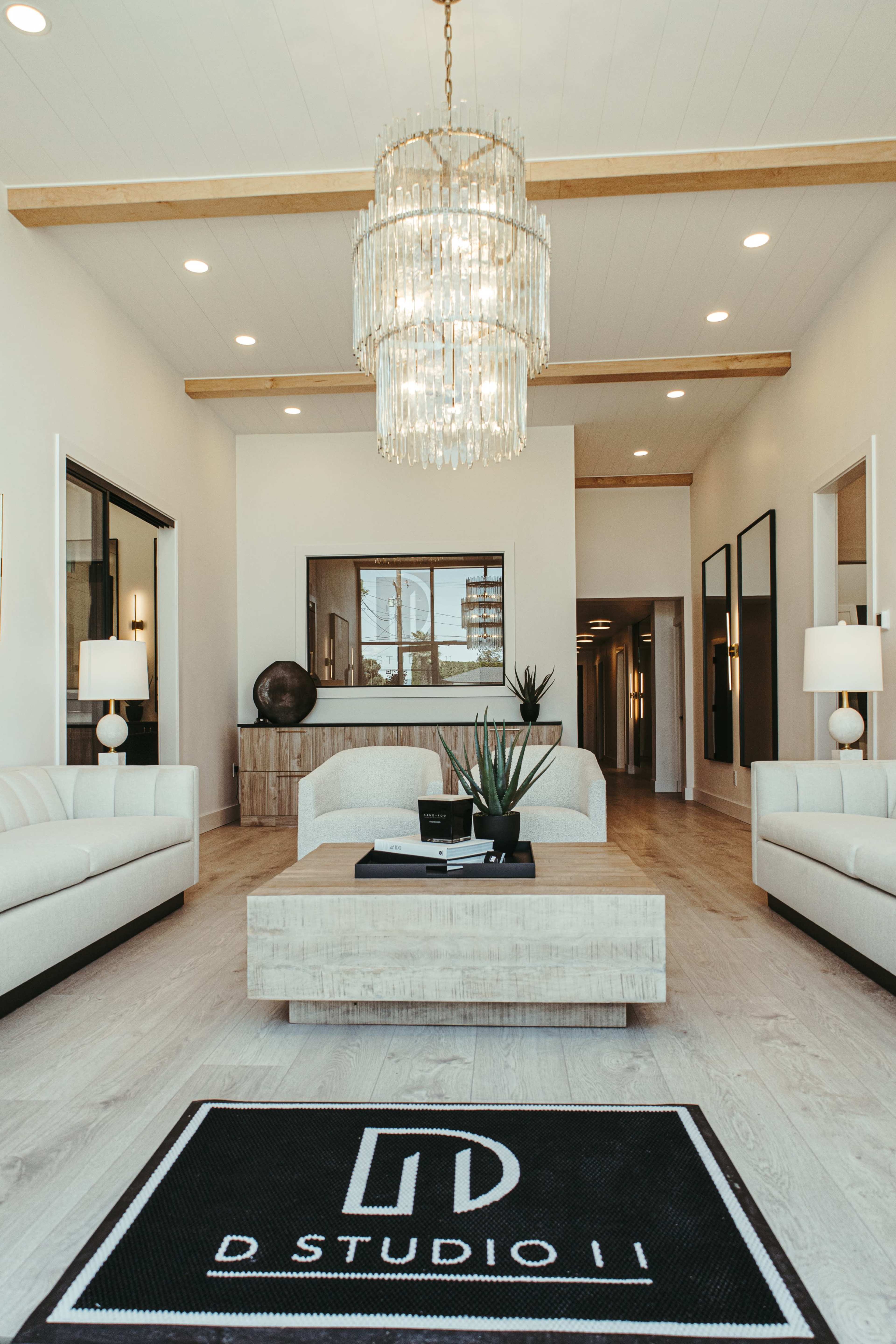 The image shows a modern interior lobby with two white sofas, a wooden coffee table, a large chandelier, and a black rug featuring the logo "D STUDIO II."