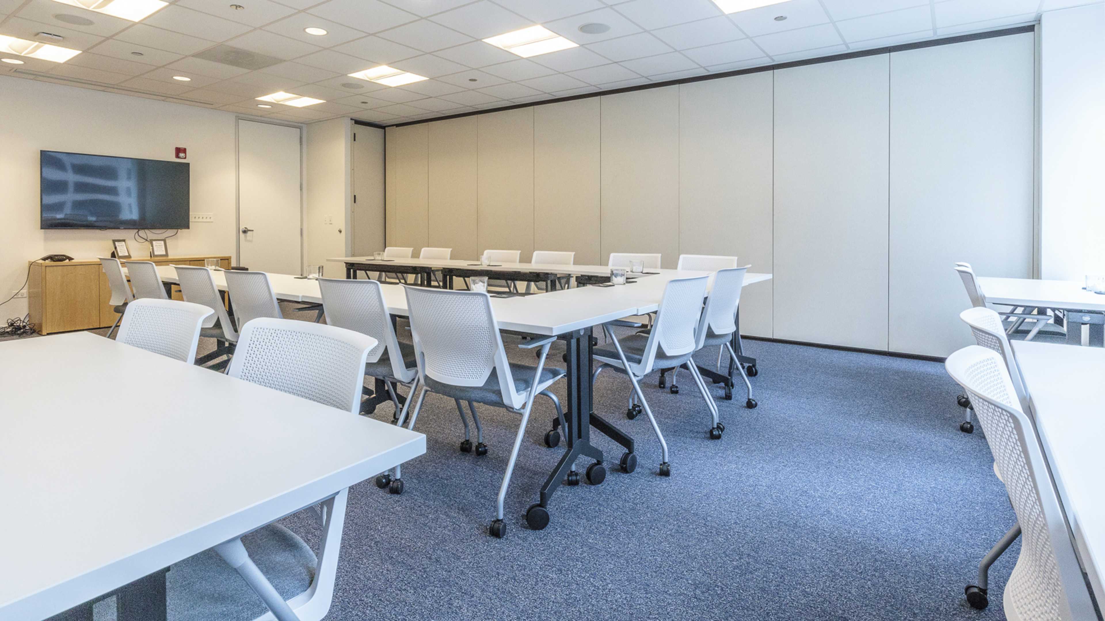 A modern conference room with multiple white tables arranged in a U-shape and several white chairs, alongside a large screen mounted on the wall.