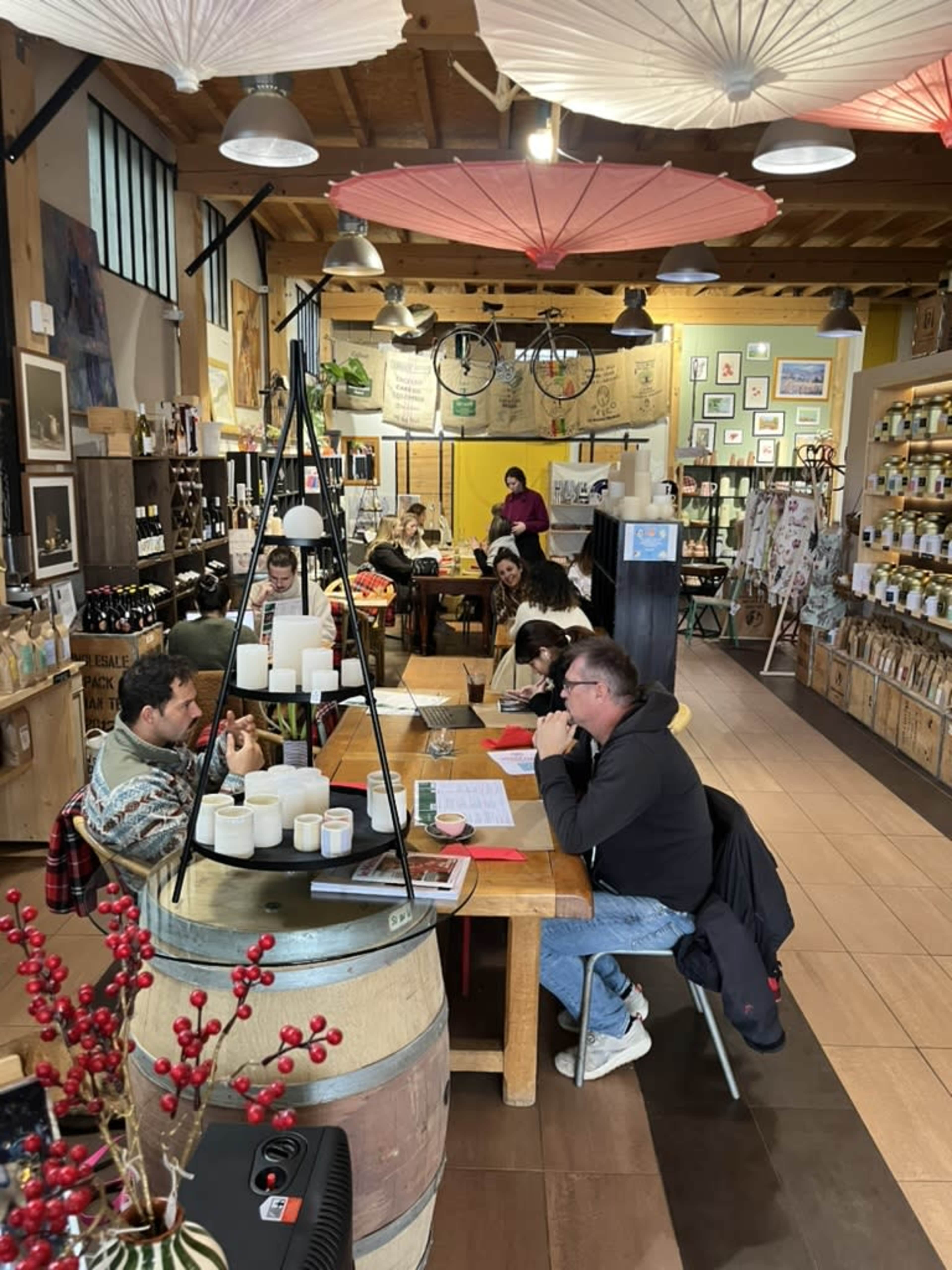 A cozy shop features wooden tables filled with patrons enjoying food and drinks, surrounded by shelves of wine and local products, and decorated with colorful paper umbrellas overhead.