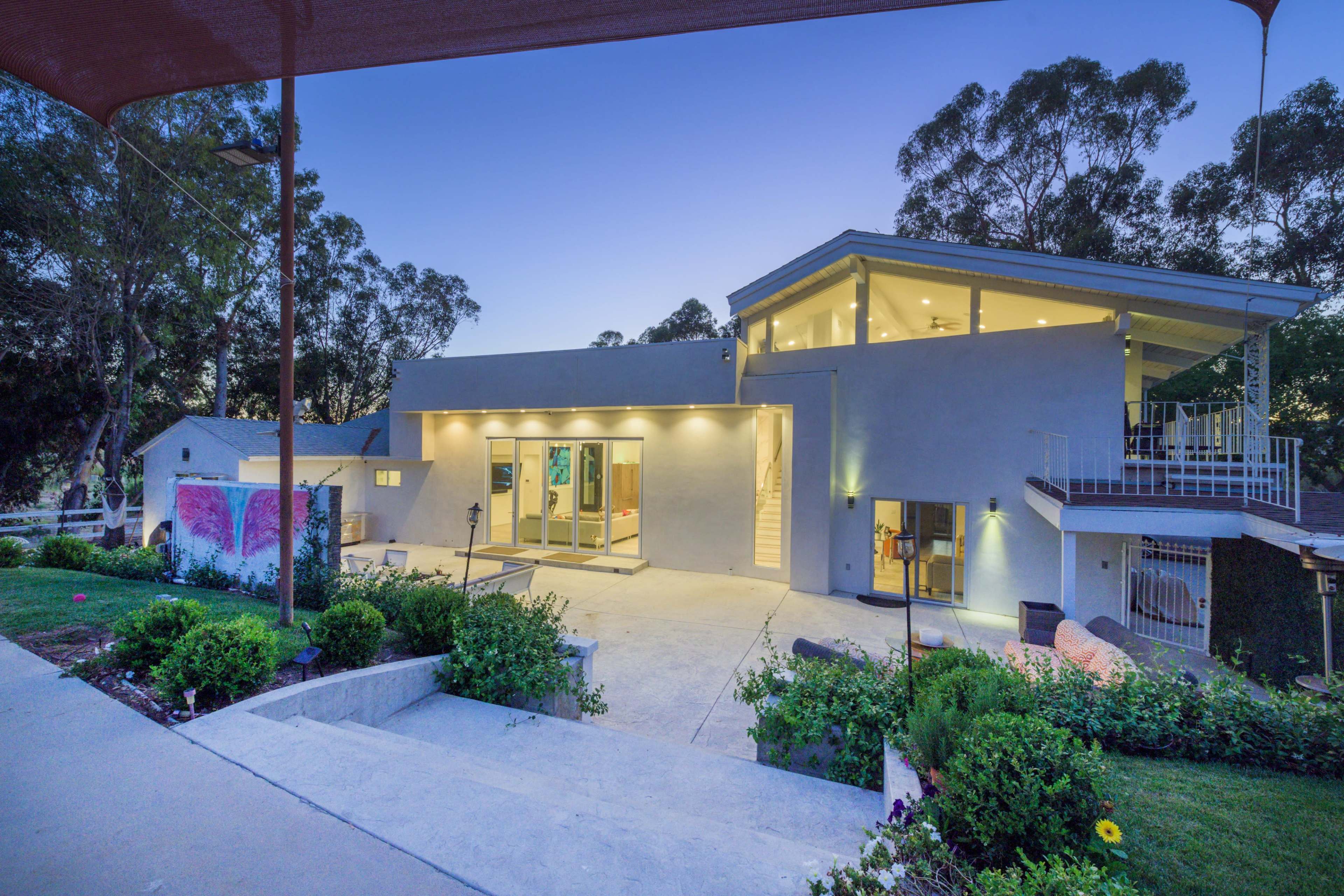 A modern two-story house with large windows, surrounded by greenery, at dusk.