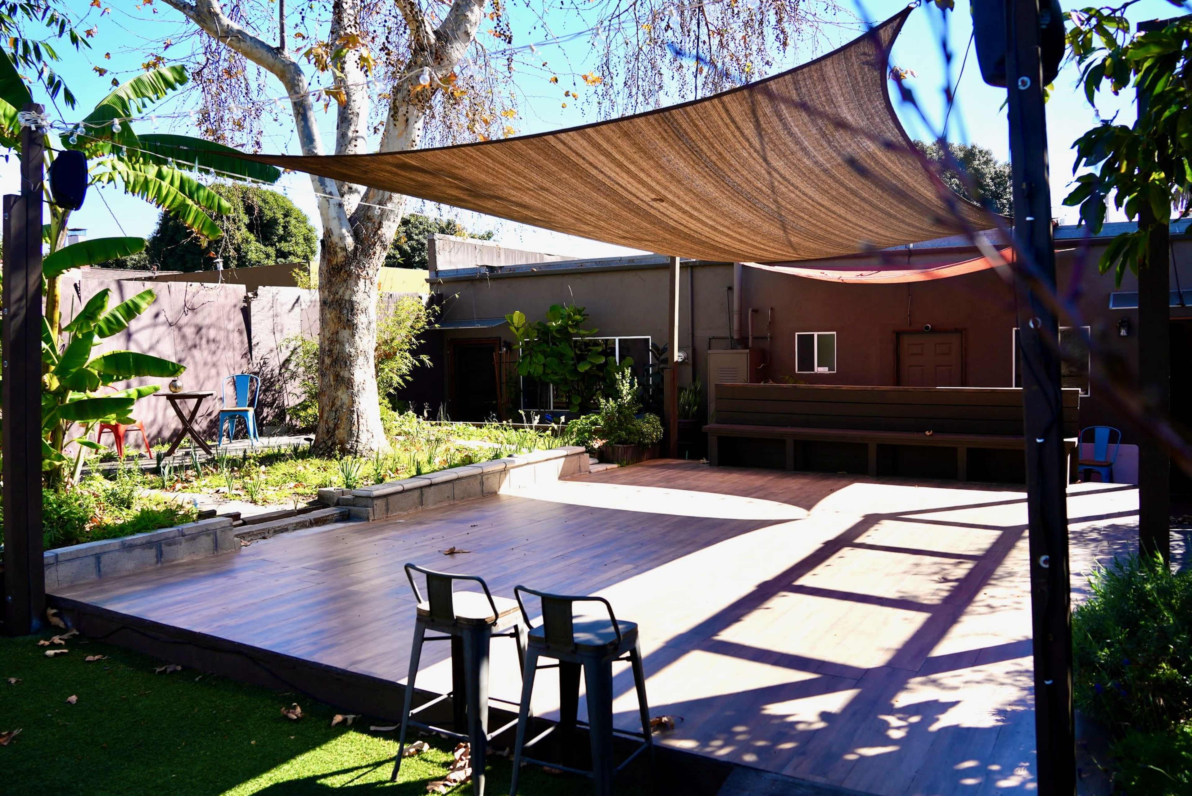 The outdoor space features a wooden deck with a shaded area covered by a large canopy, surrounded by greenery and a few seating options.