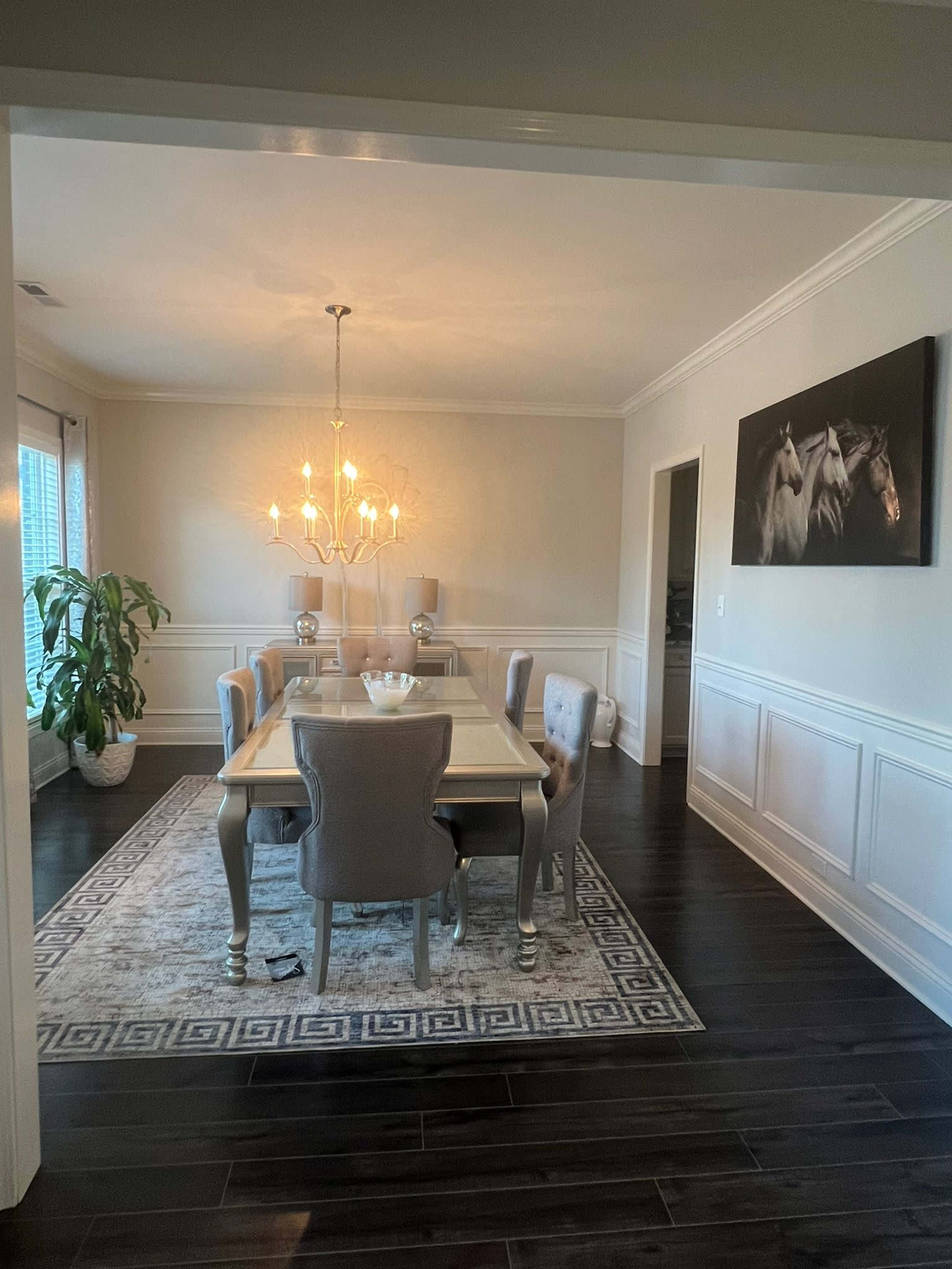 A spacious dining room features a large table surrounded by upholstered chairs, a chandelier above, and a decorative rug on the dark wooden floor.