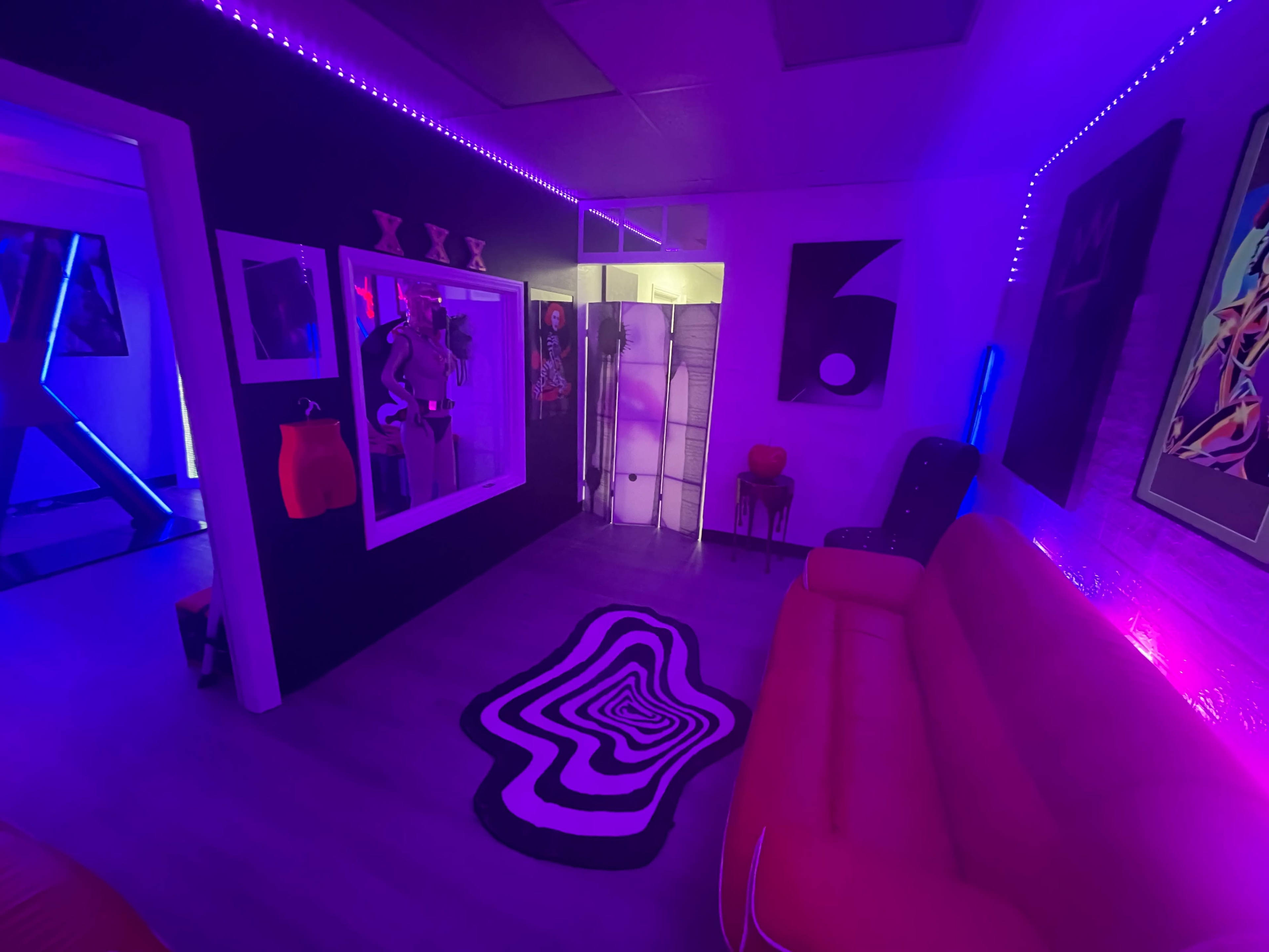 A colorful lounge area with neon lighting, a black and white patterned rug, a red couch, and a mirror displaying mannequins in an artistic setting.
