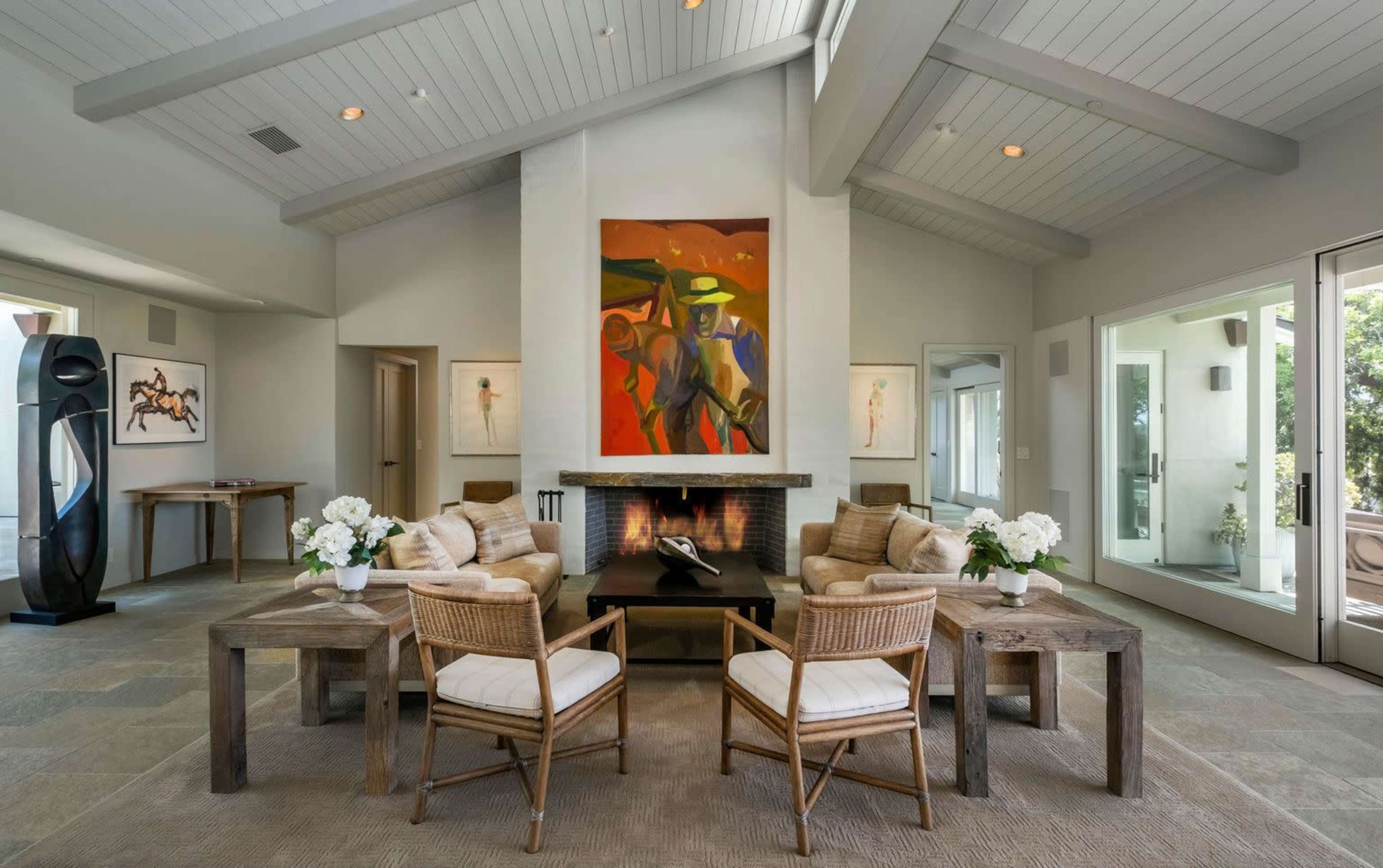 A modern living room featuring two armchairs and a coffee table arranged around a fireplace, with a large abstract painting on the wall and large windows allowing natural light.