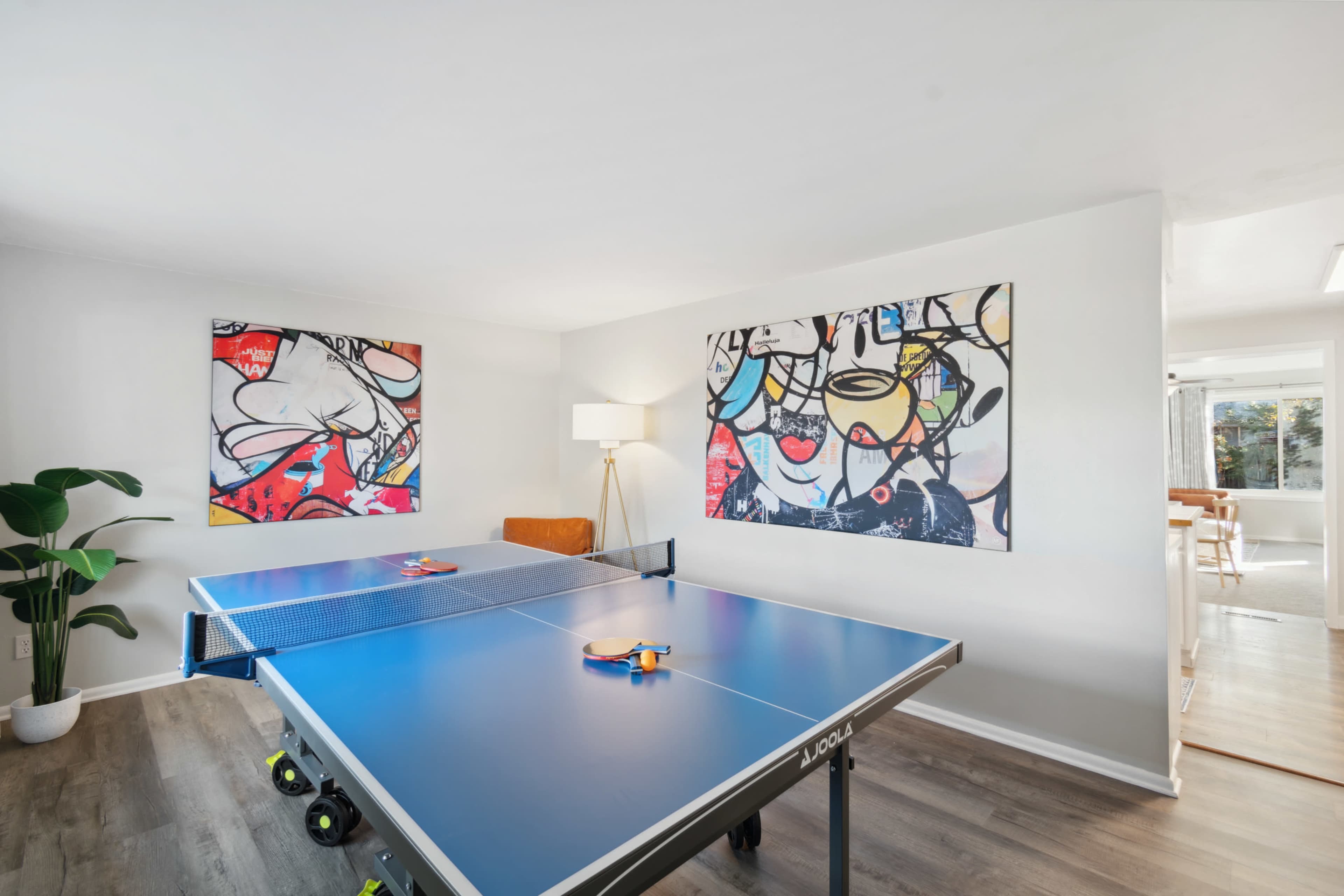 A bright room with a blue ping pong table in the center, surrounded by abstract art on the walls and a lamp beside the table.