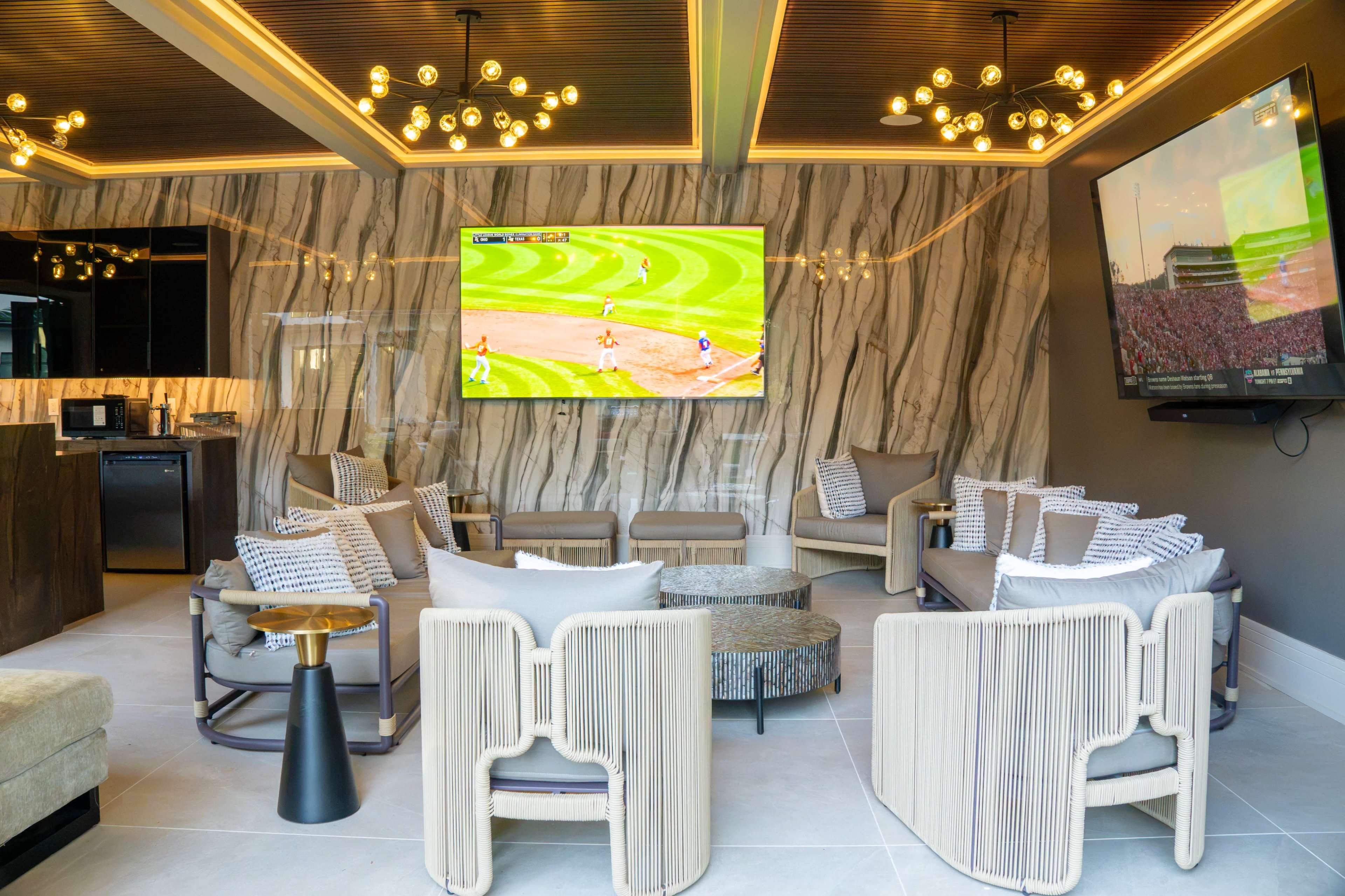 The image shows a modern lounge area with a large flat-screen television displaying a sports game, surrounded by stylish seating and decorative lighting against a marble wall backdrop.