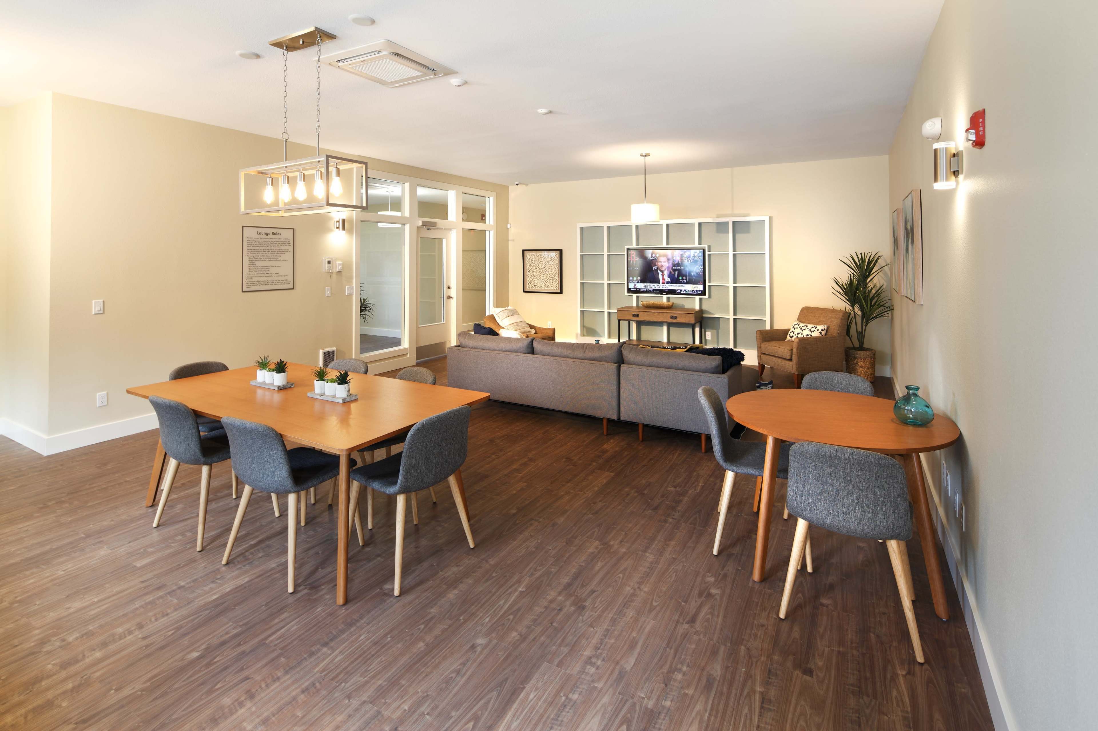 A modern communal lounge area with two dining tables, a couch, a television, and large windows letting in natural light.