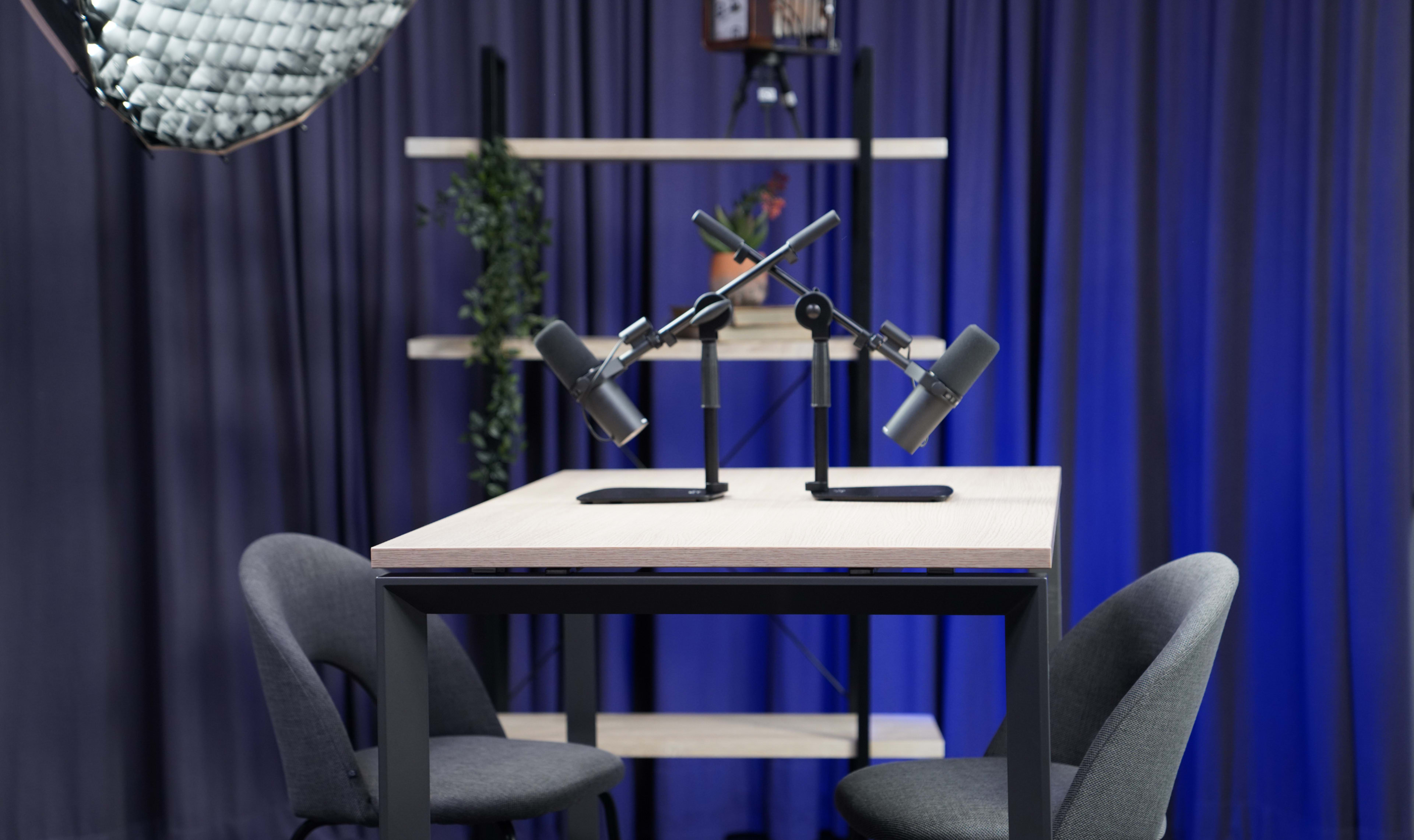 The image shows a modern, minimalist recording studio setup with two microphones positioned on a table, flanked by two chairs and background shelves.
