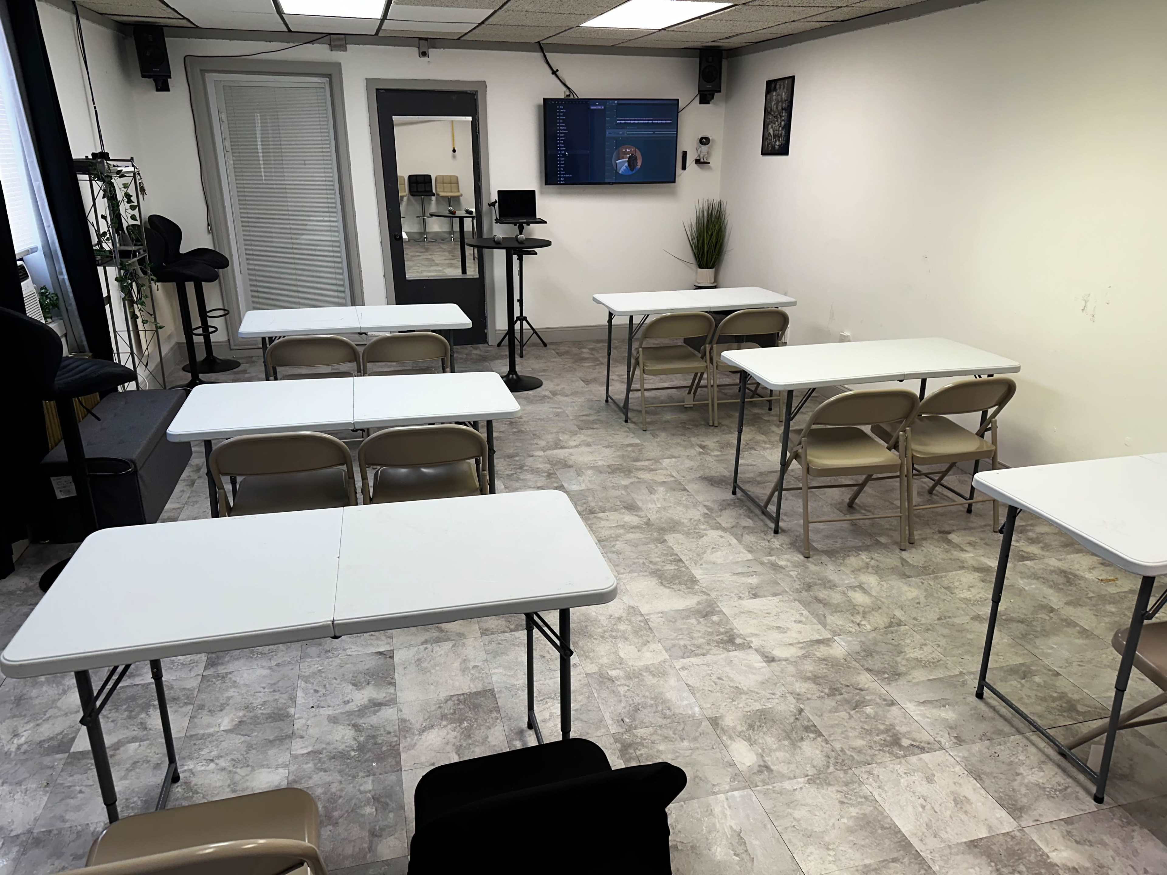 A classroom features six white tables arranged with folding chairs, and a large screen displaying content in the background.