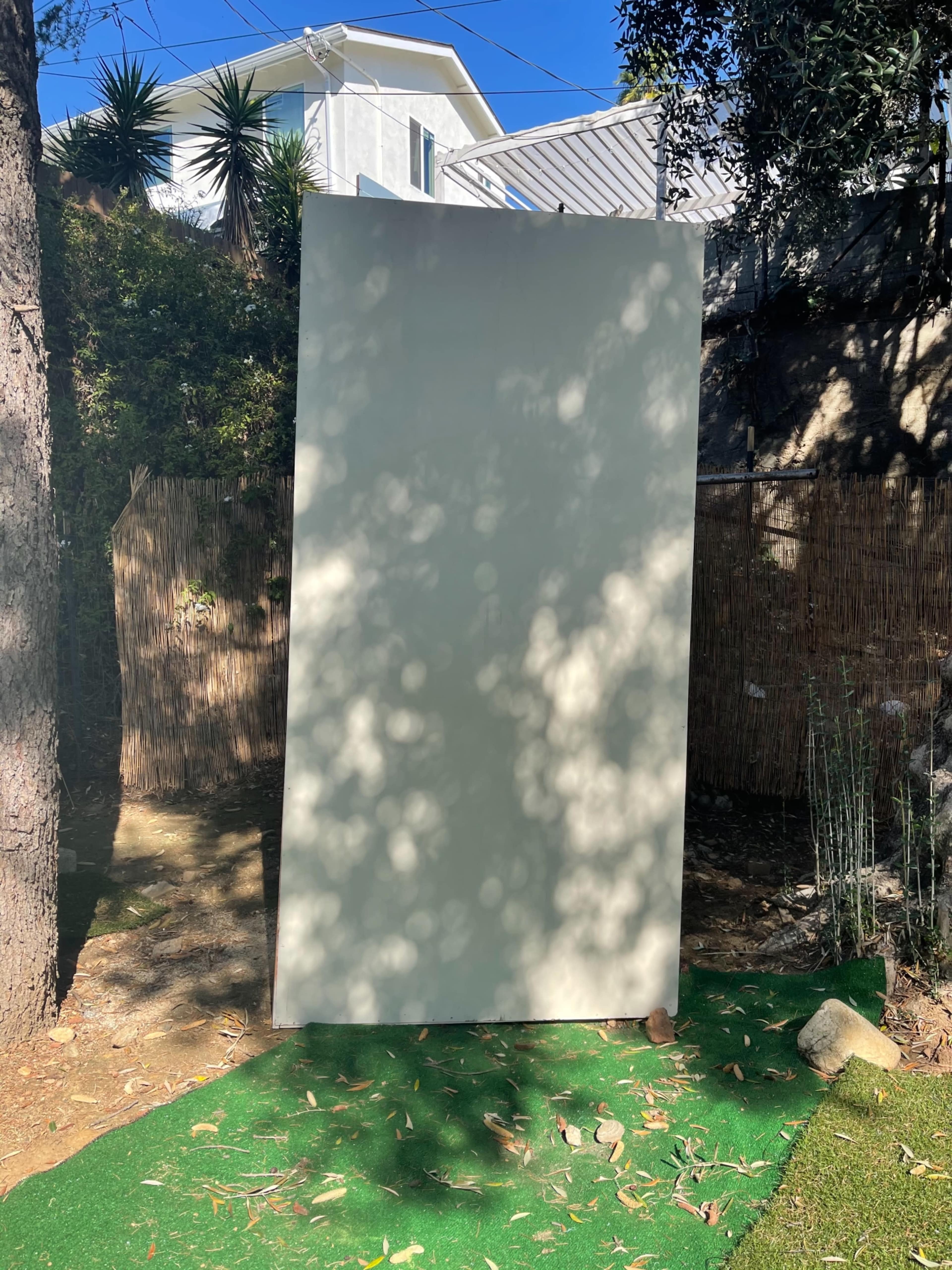 A large blank canvas stands upright on a grassy patch surrounded by trees and bamboo fencing.