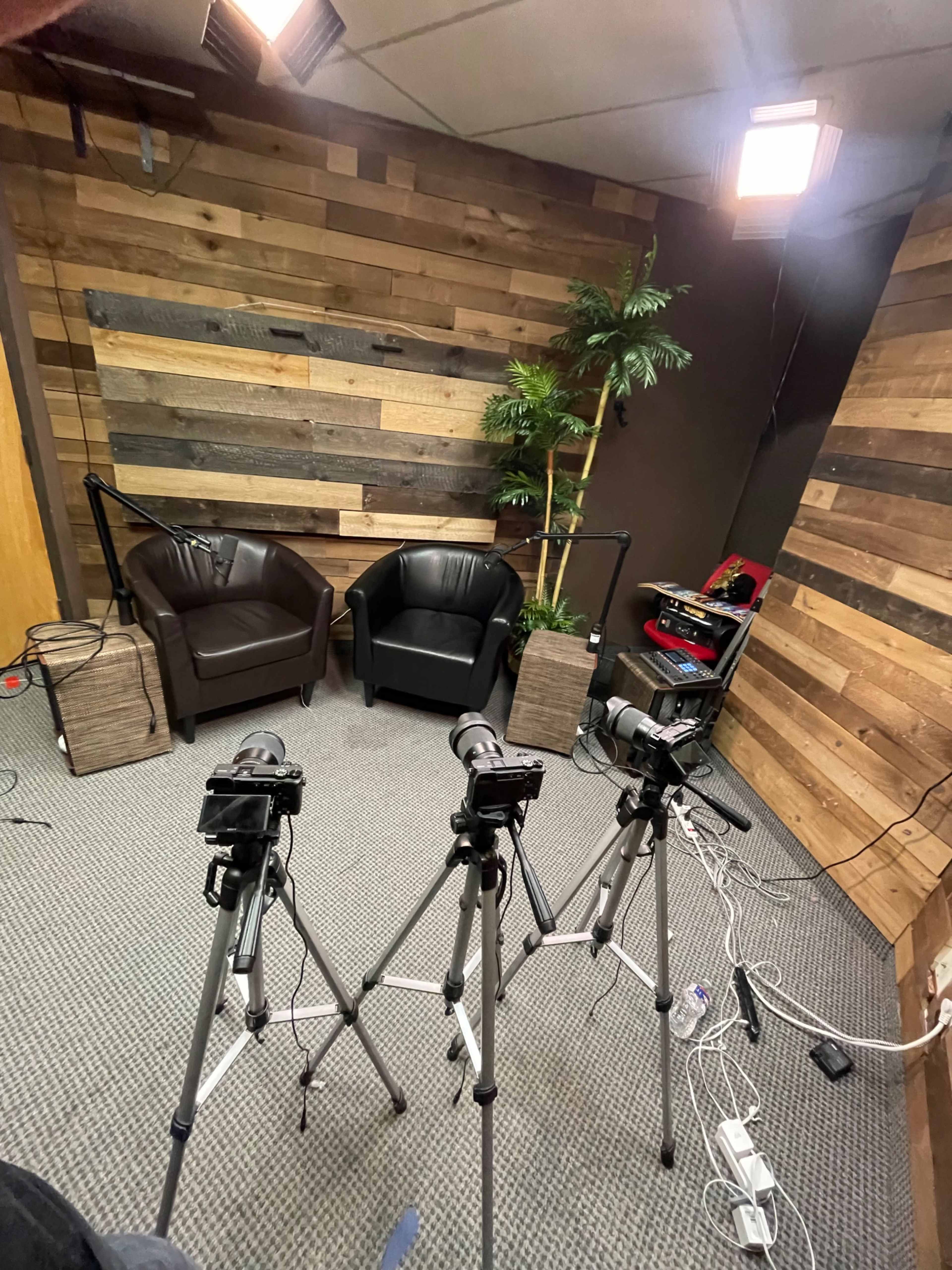 A videography setup with three cameras on tripods facing two black chairs in a room featuring wooden paneling and a potted plant.