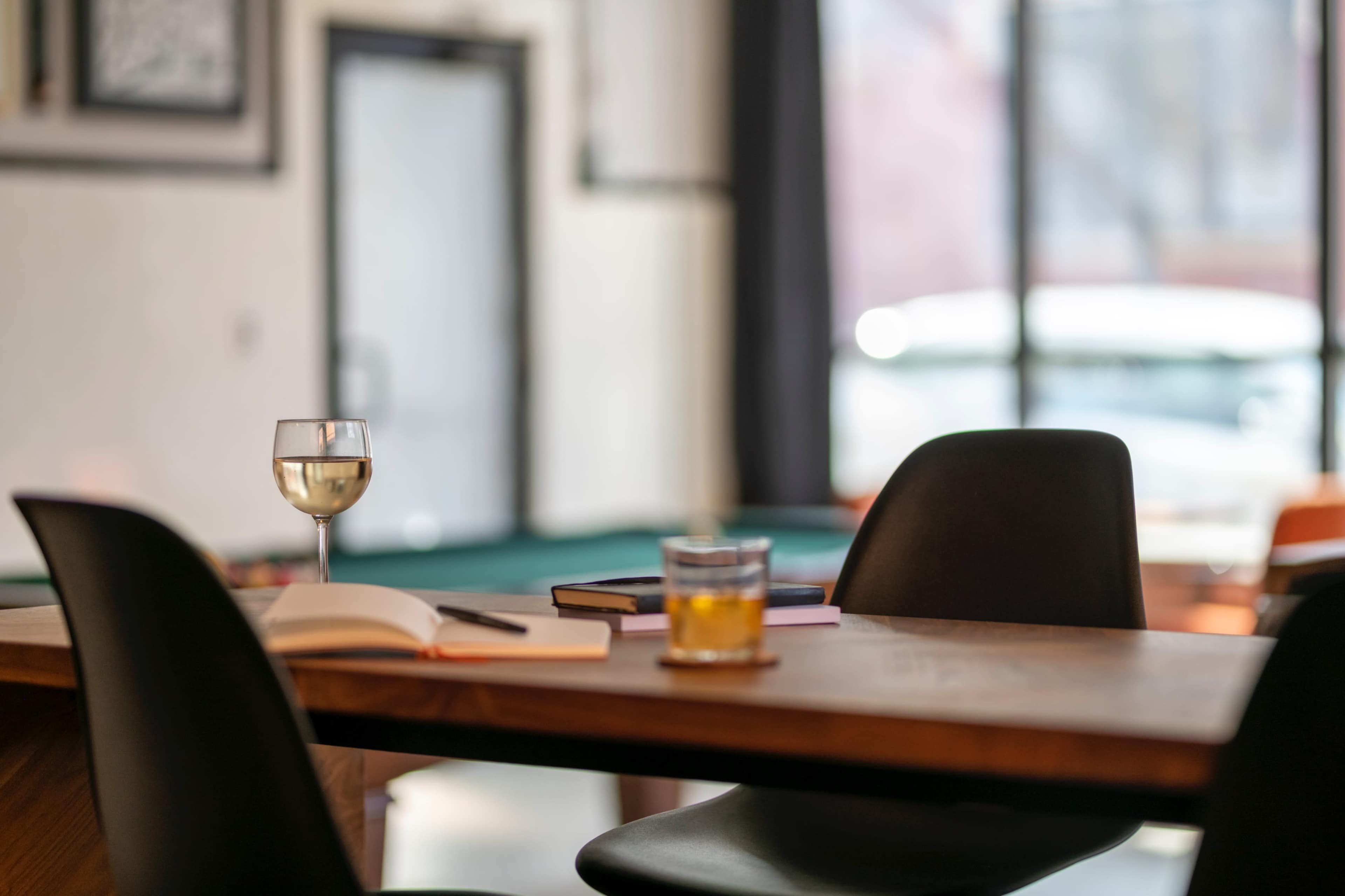 A wooden table in a relaxed setting holds a glass of wine, a rocks glass, and a few books, with chairs positioned around it.