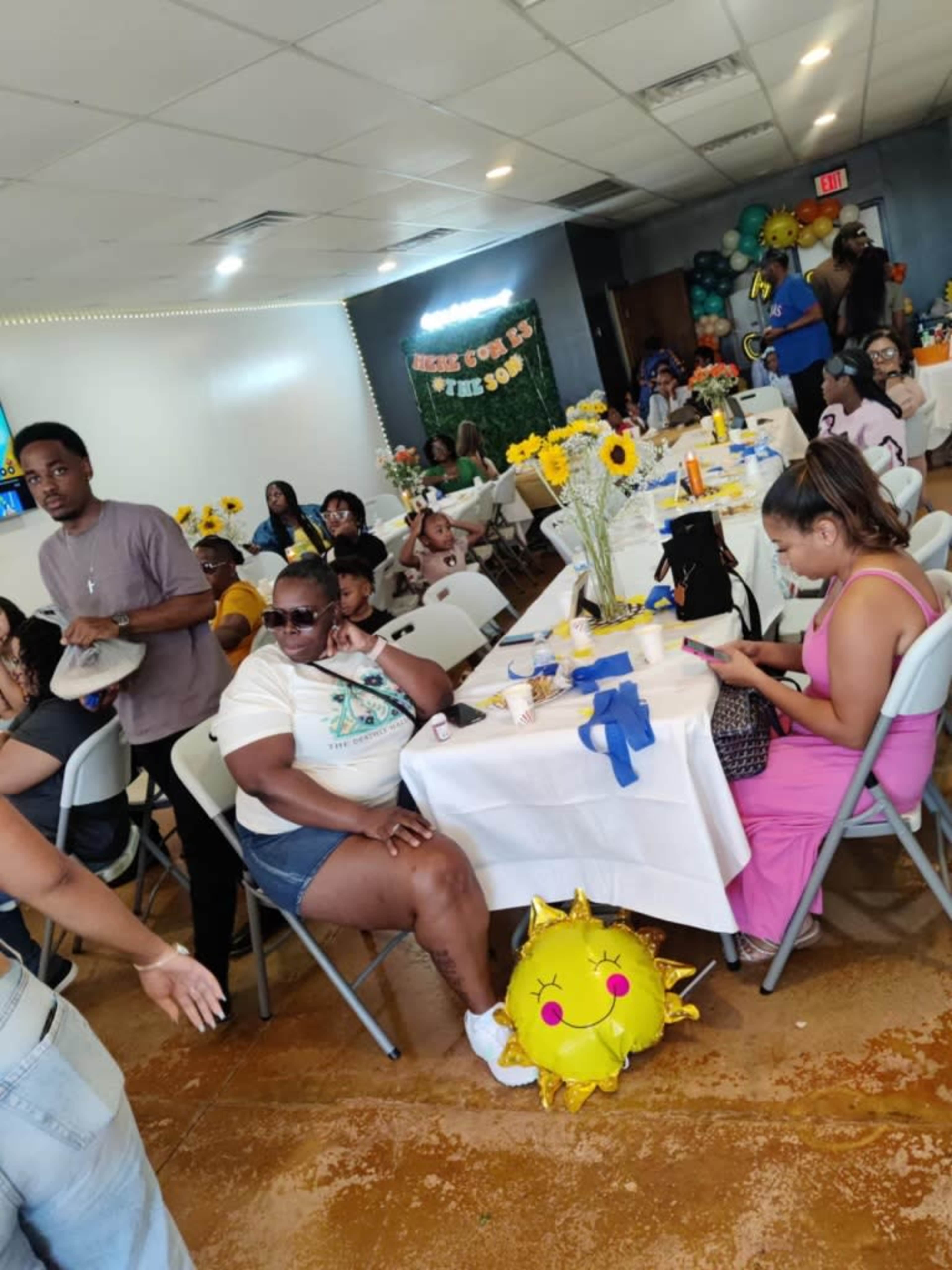 A diverse group of people sits at tables decorated with flowers and balloons in a bright event space.