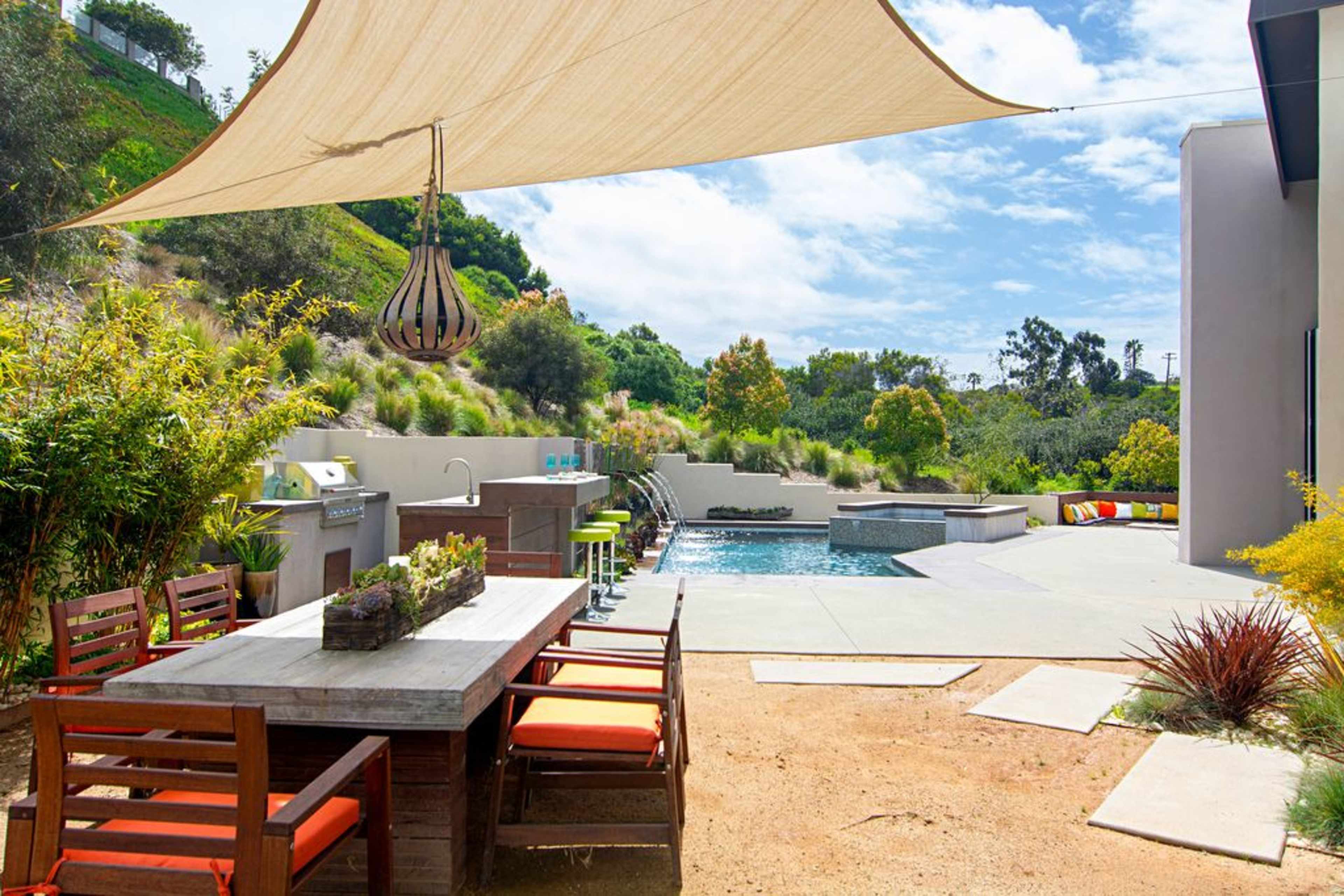A modern outdoor area featuring a dining table, a pool with a spa, and a sunshade above, surrounded by greenery.