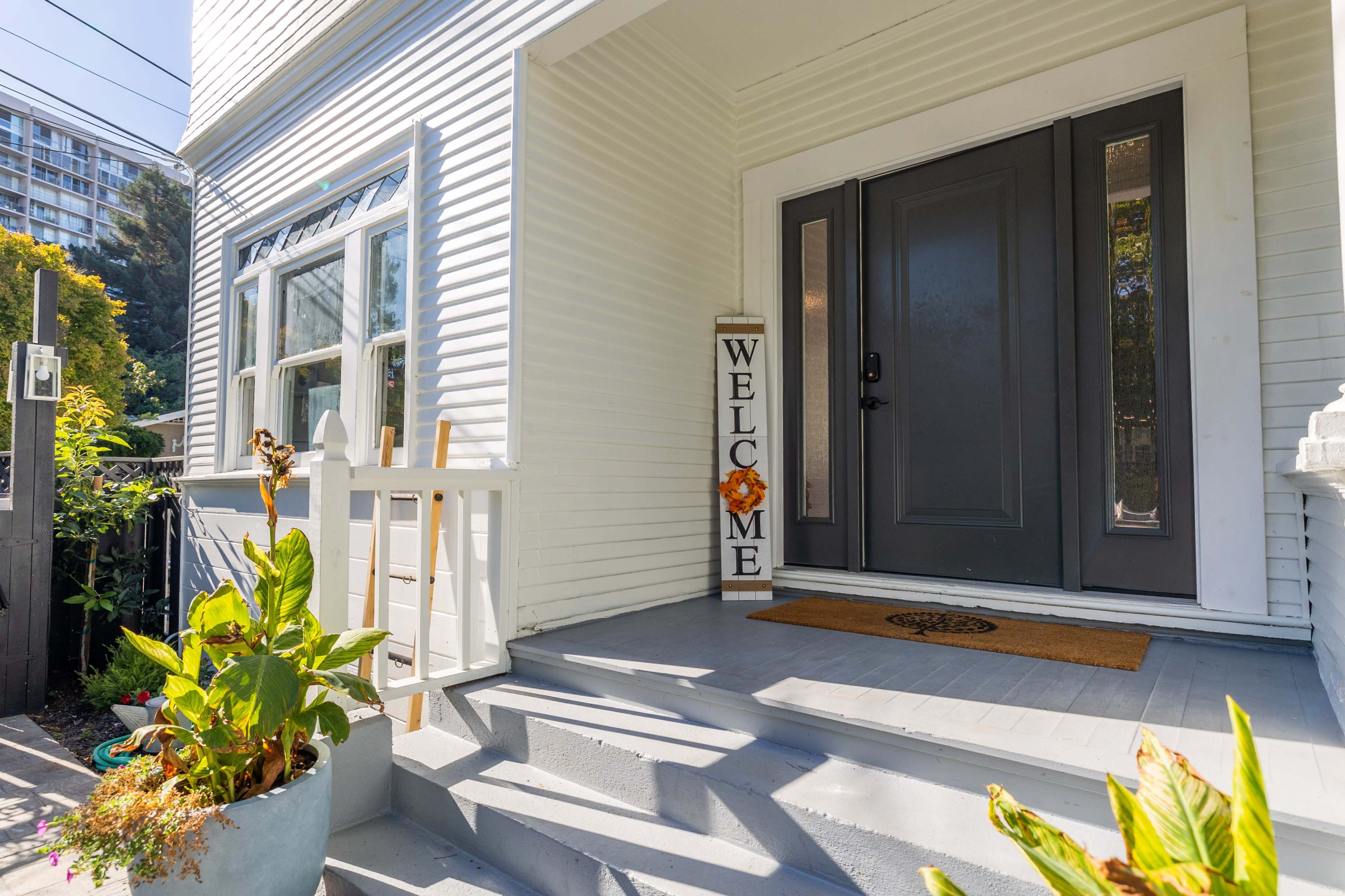 A front porch with a dark front door, a welcome sign by the entrance, and potted plants on the steps.