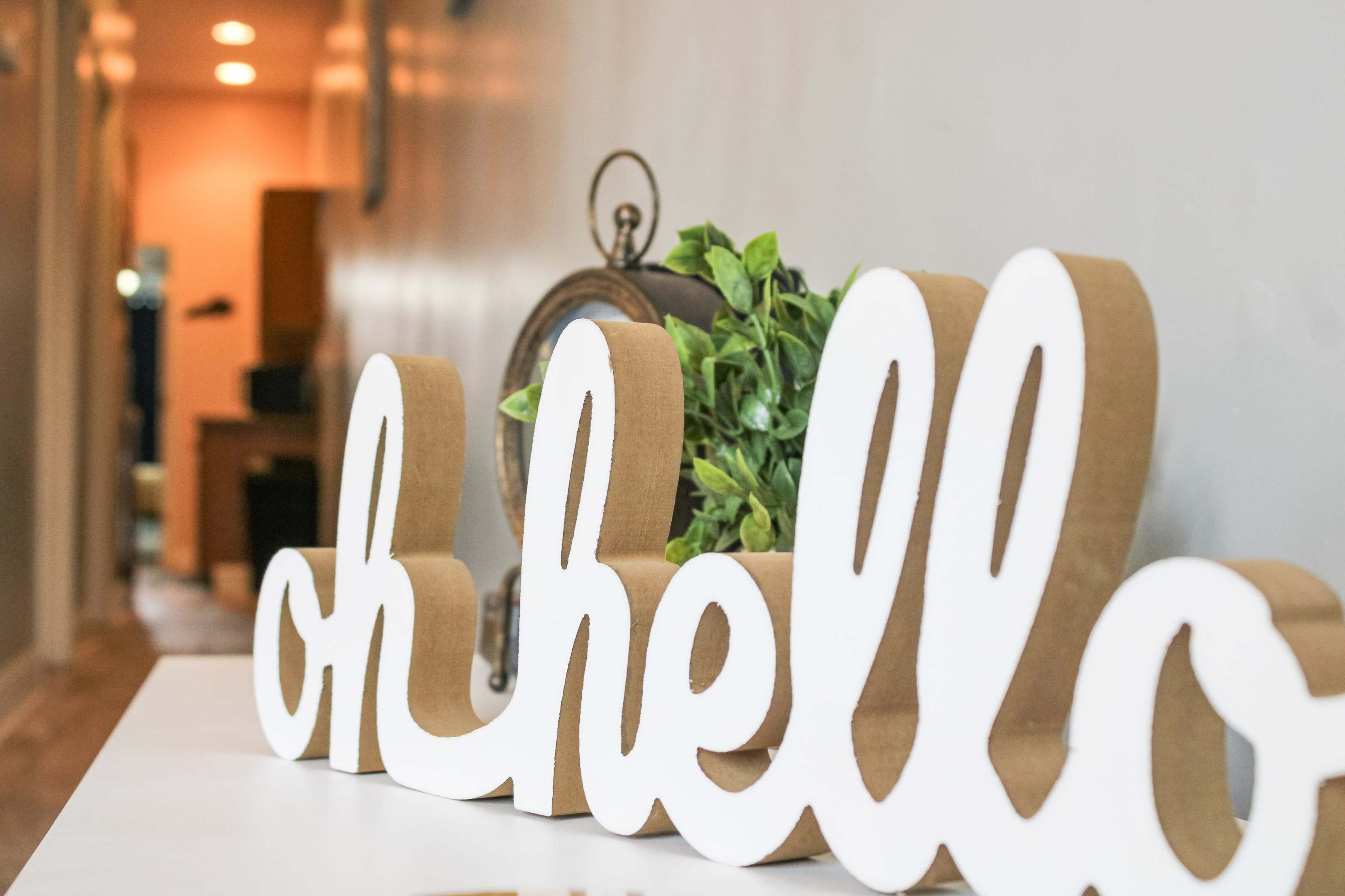 A decorative "oh hello" wooden sign sits on a table, with a green plant nearby, while a hallway leads to softly illuminated rooms in the background.
