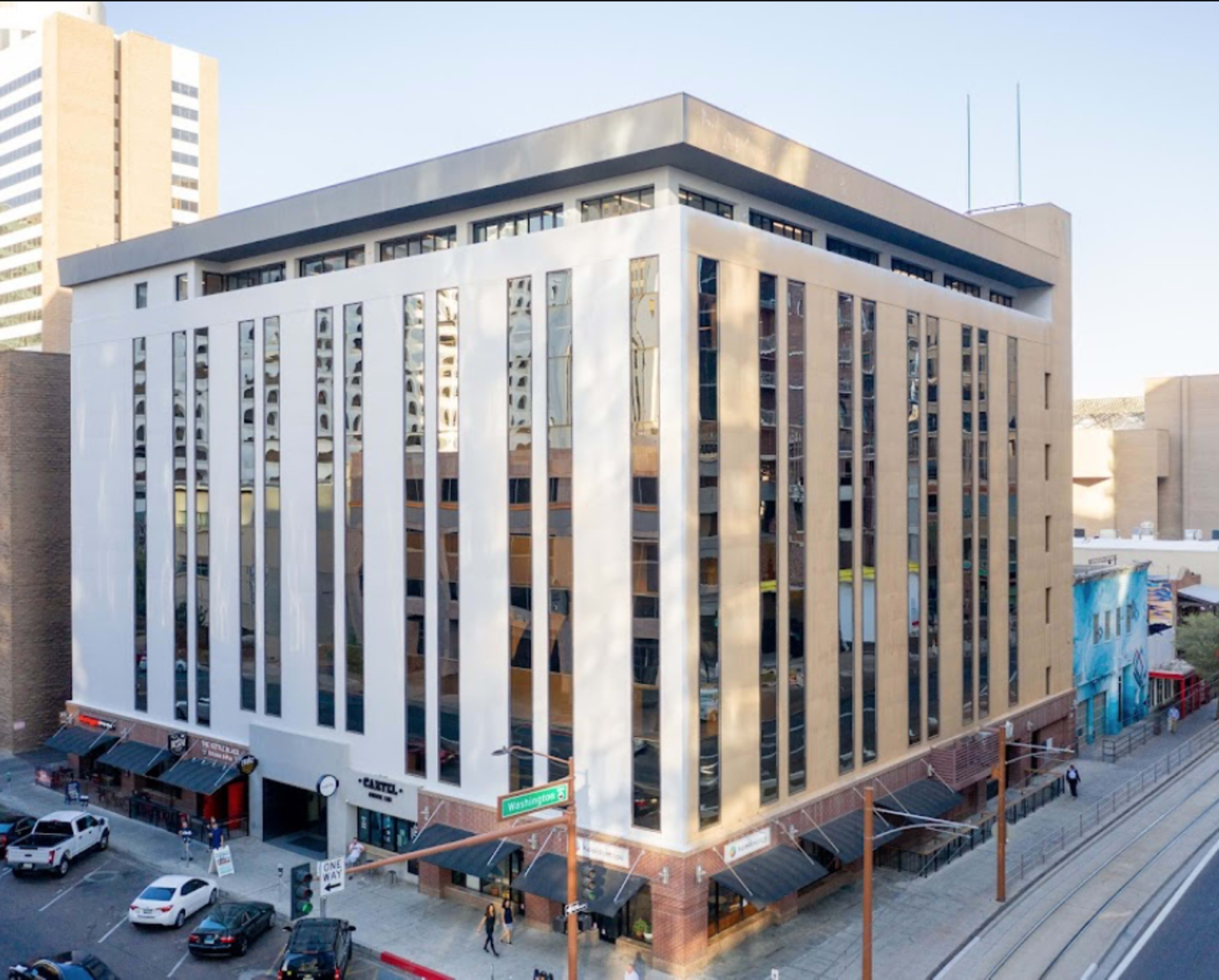 A modern multi-story building with large glass windows is located at a street intersection, flanked by parked cars and adjacent businesses.