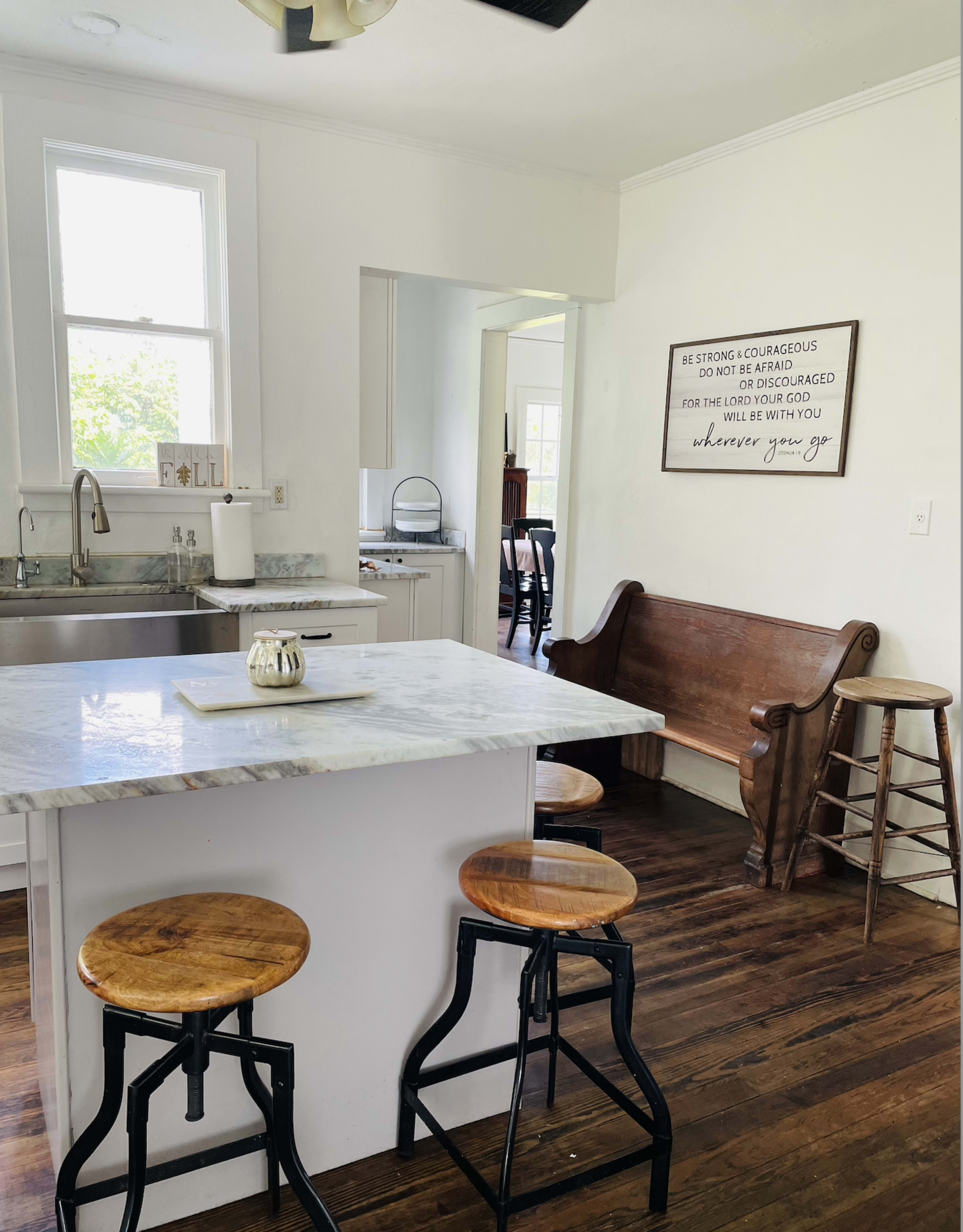 The image shows a bright kitchen with a marble countertop island, two wooden stools, and a rustic bench against a wall featuring a decorative sign.