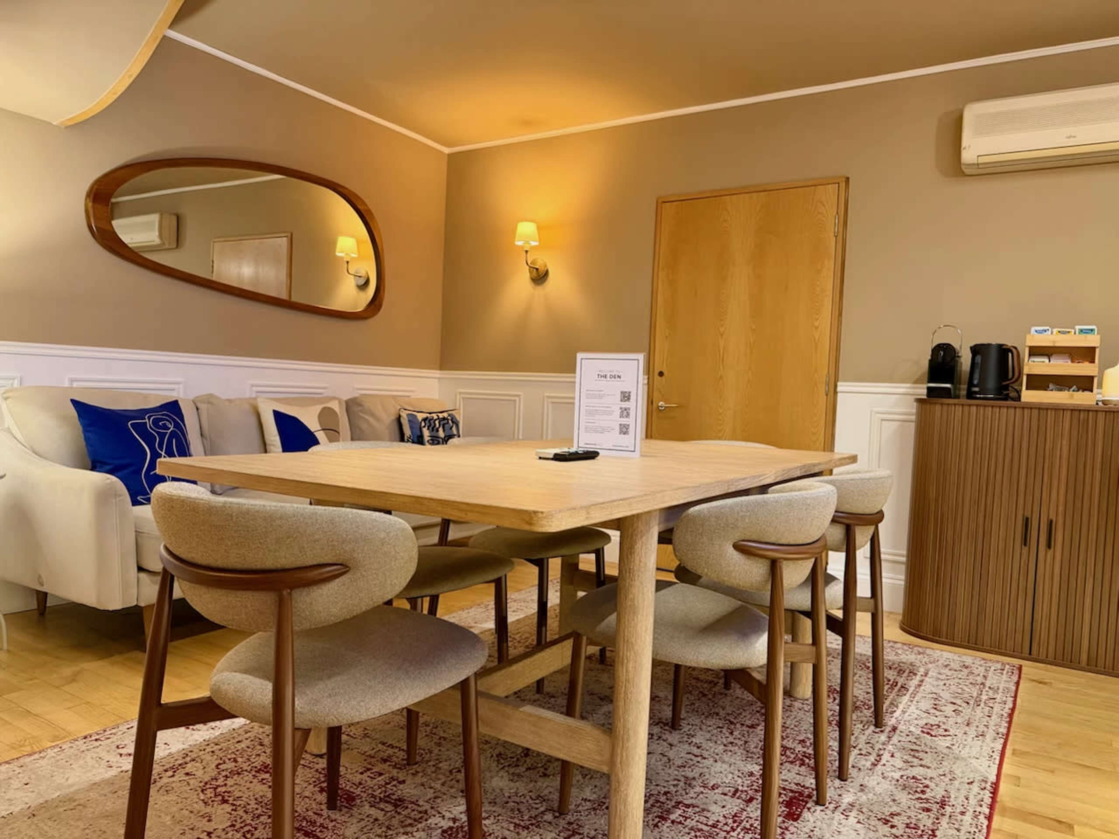 The image shows a modern dining area with a wooden table surrounded by six upholstered chairs, a beige sofa, and light-colored walls, along with a mirror and a wooden cabinet in the background.
