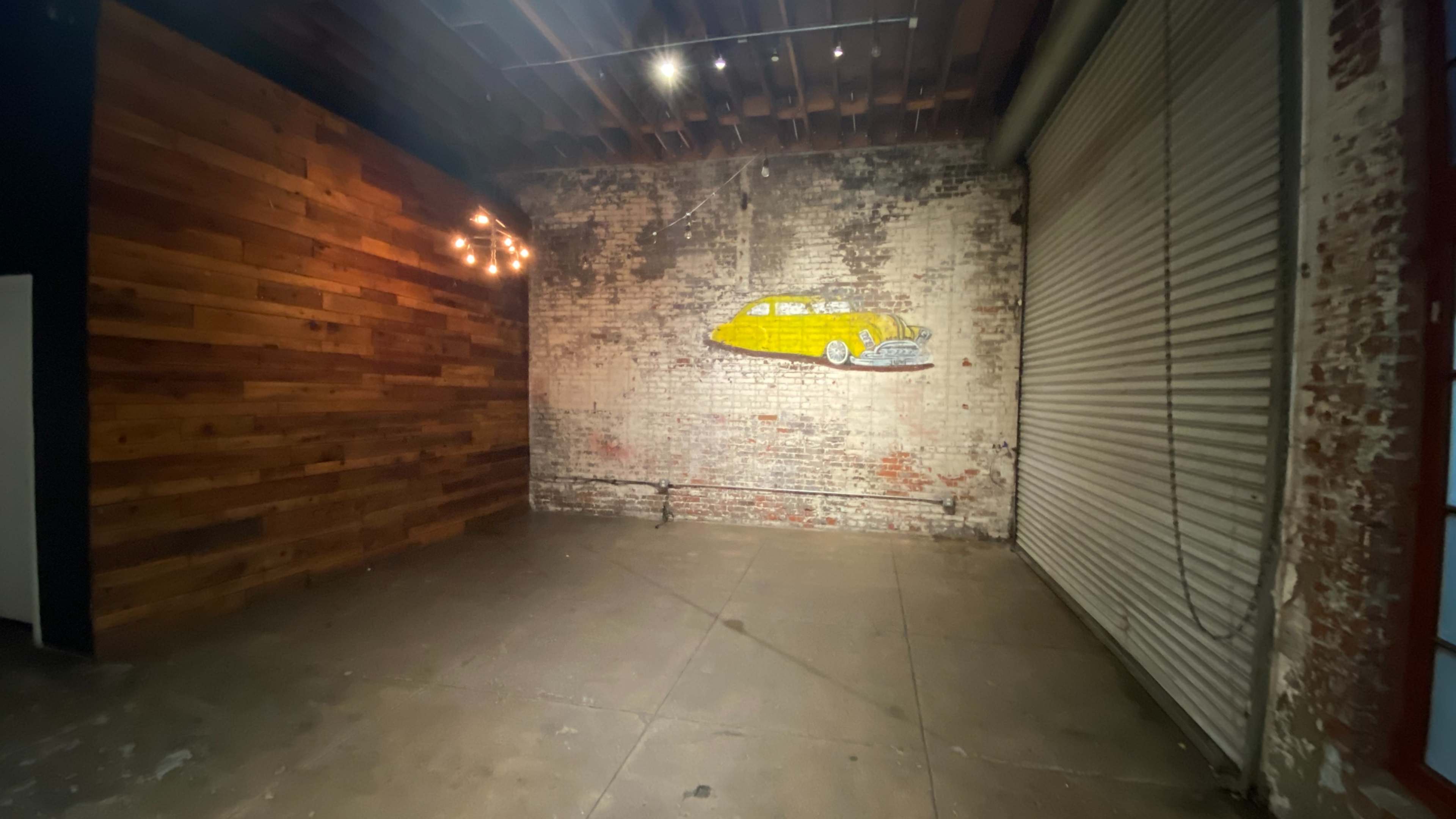 An empty industrial space with a brick wall, wooden paneling, and a mural of a yellow surfboard beside a large shutter door.