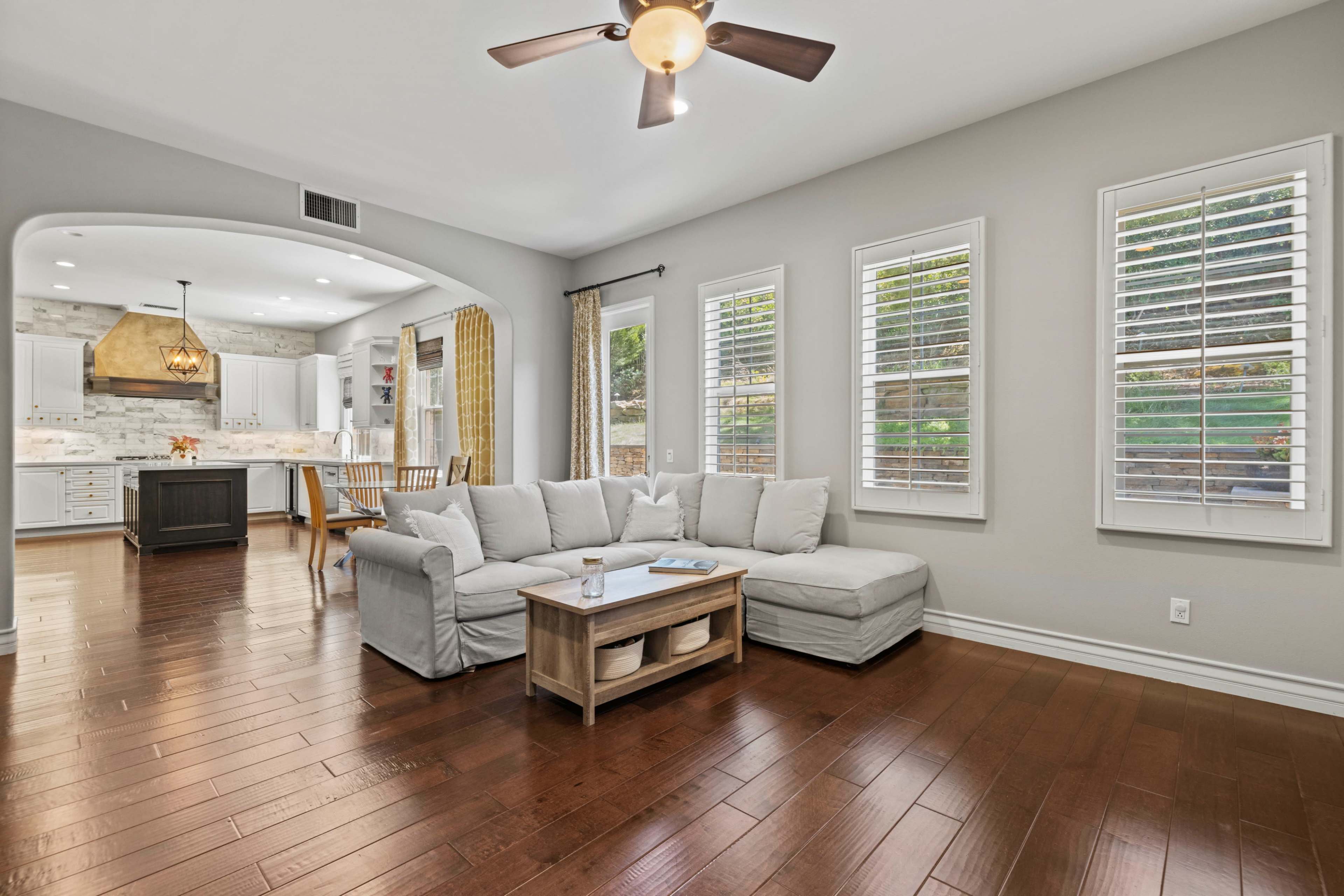 A living room with a light-colored sectional sofa, wooden flooring, and large windows letting in natural light, leading into a kitchen area with a pendant light.