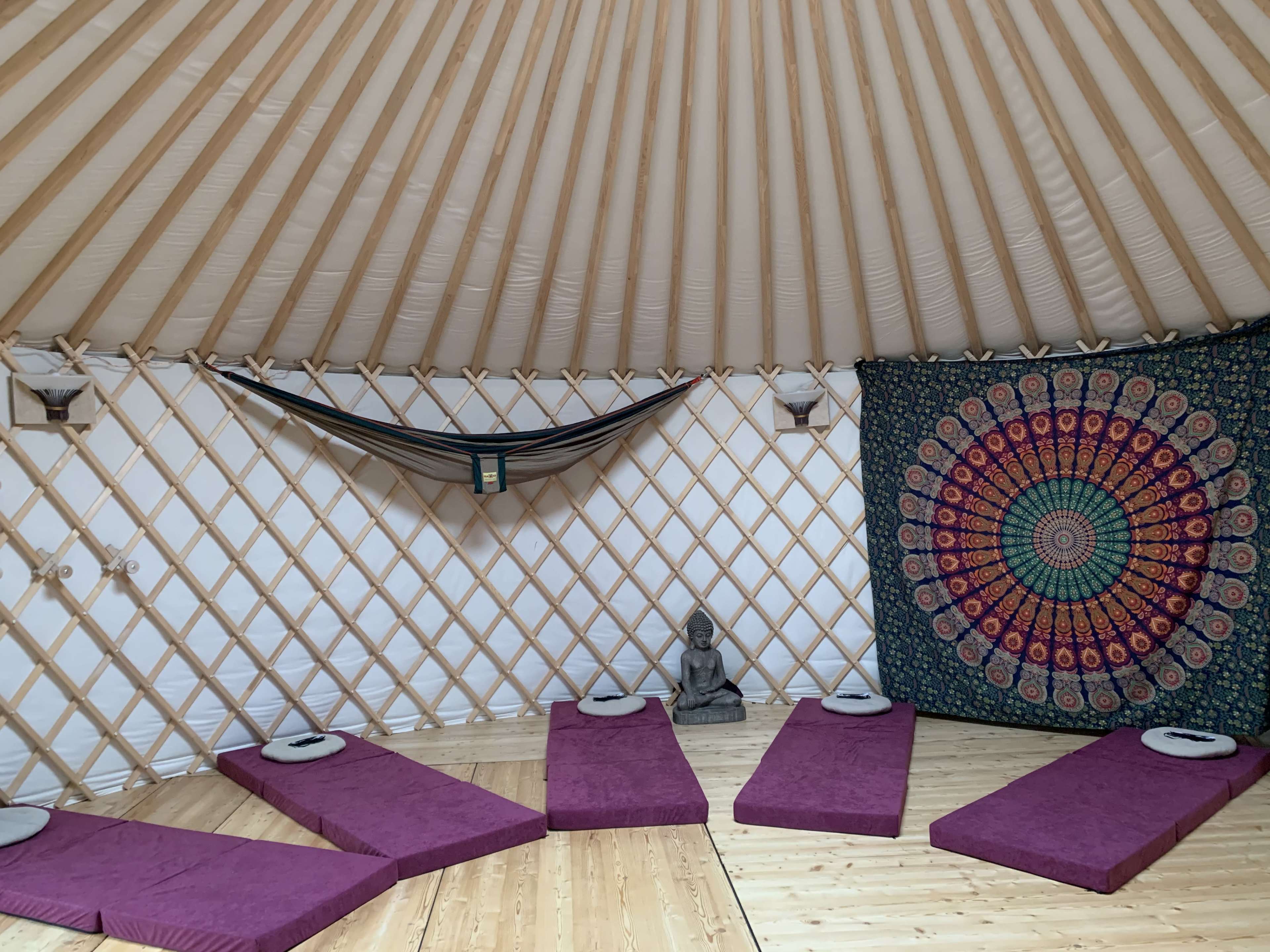 The interior of a yurt features purple mats on a wooden floor, a central statue of a Buddha, a colorful tapestry, and a hammock hanging on the wall.