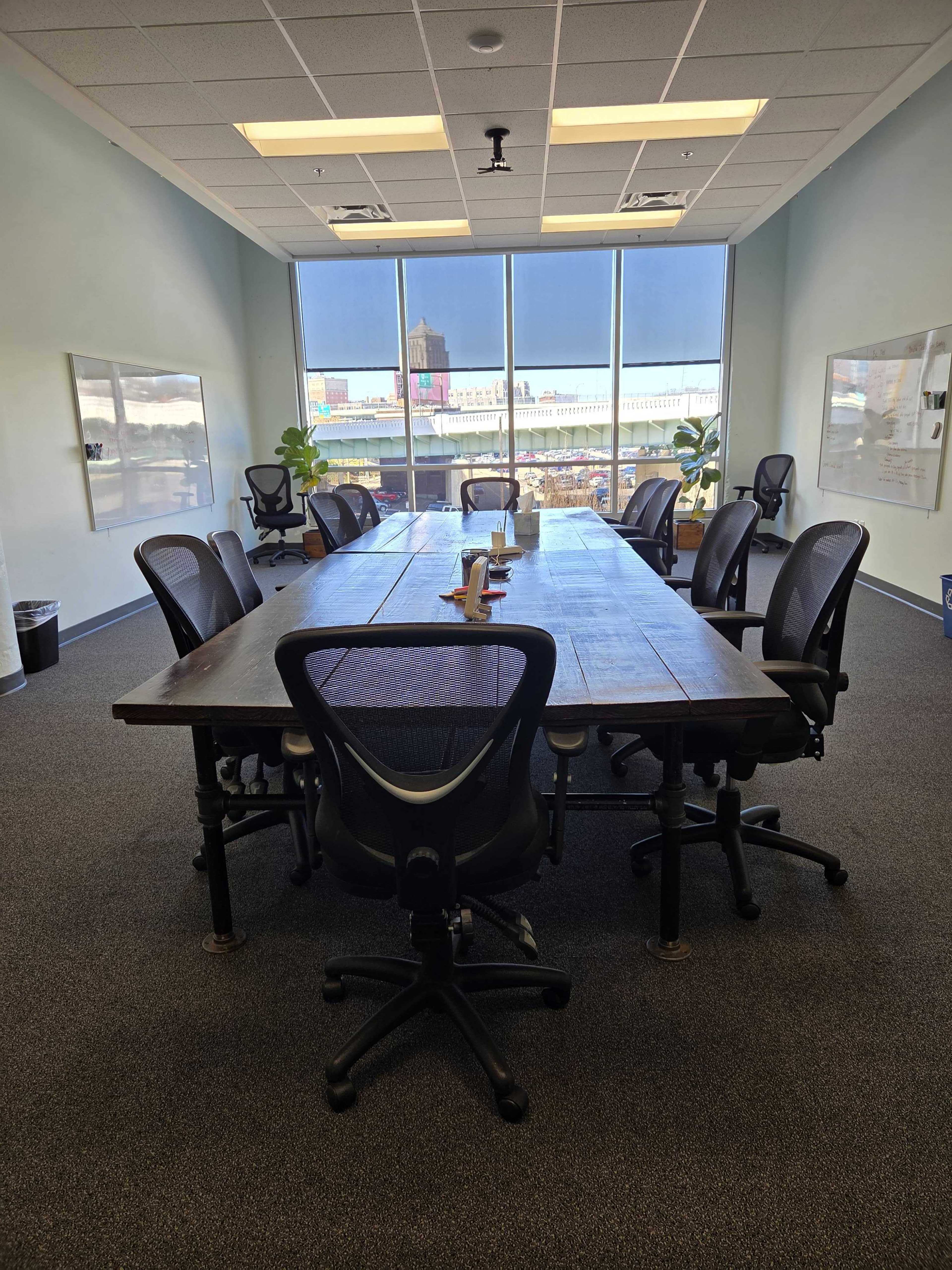A large, rectangular conference table is surrounded by several ergonomic chairs in a well-lit meeting room with windows that show a view of a cityscape.