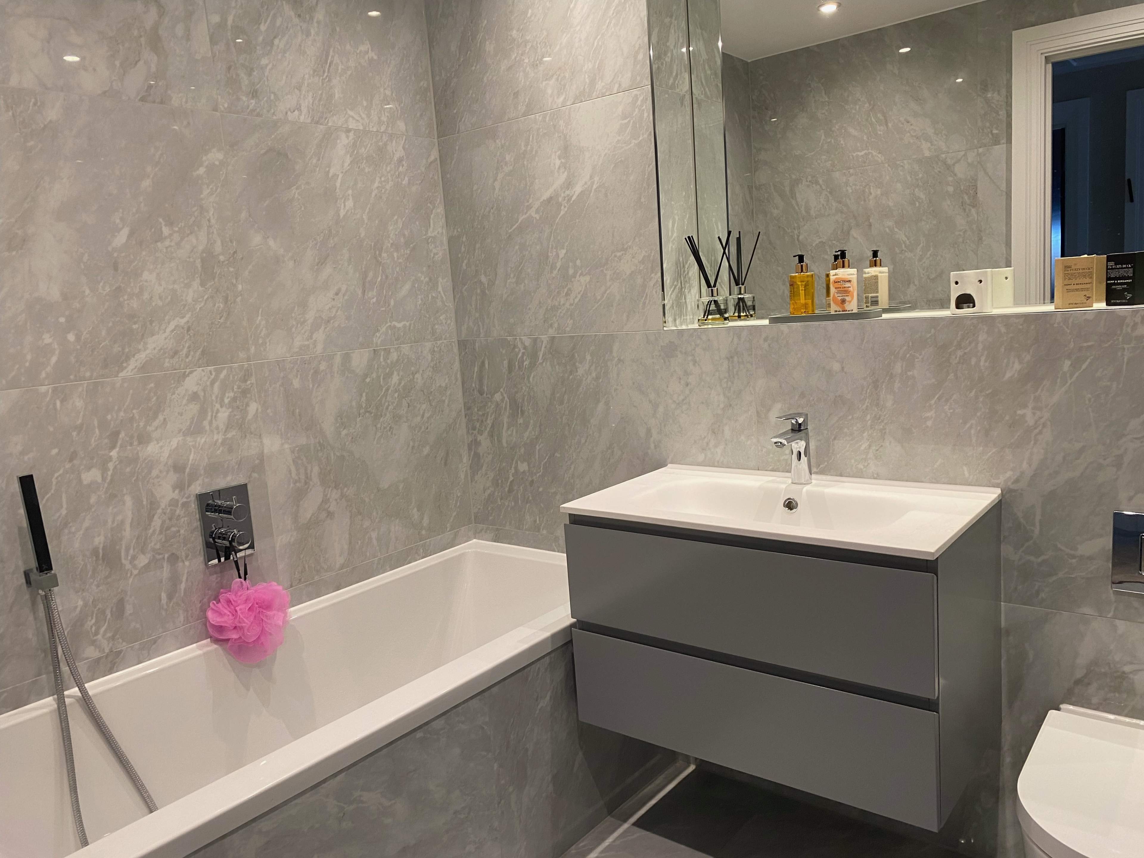 A modern bathroom with gray marble walls, a bathtub, a sink with a cabinet below, and various toiletries on the counter.