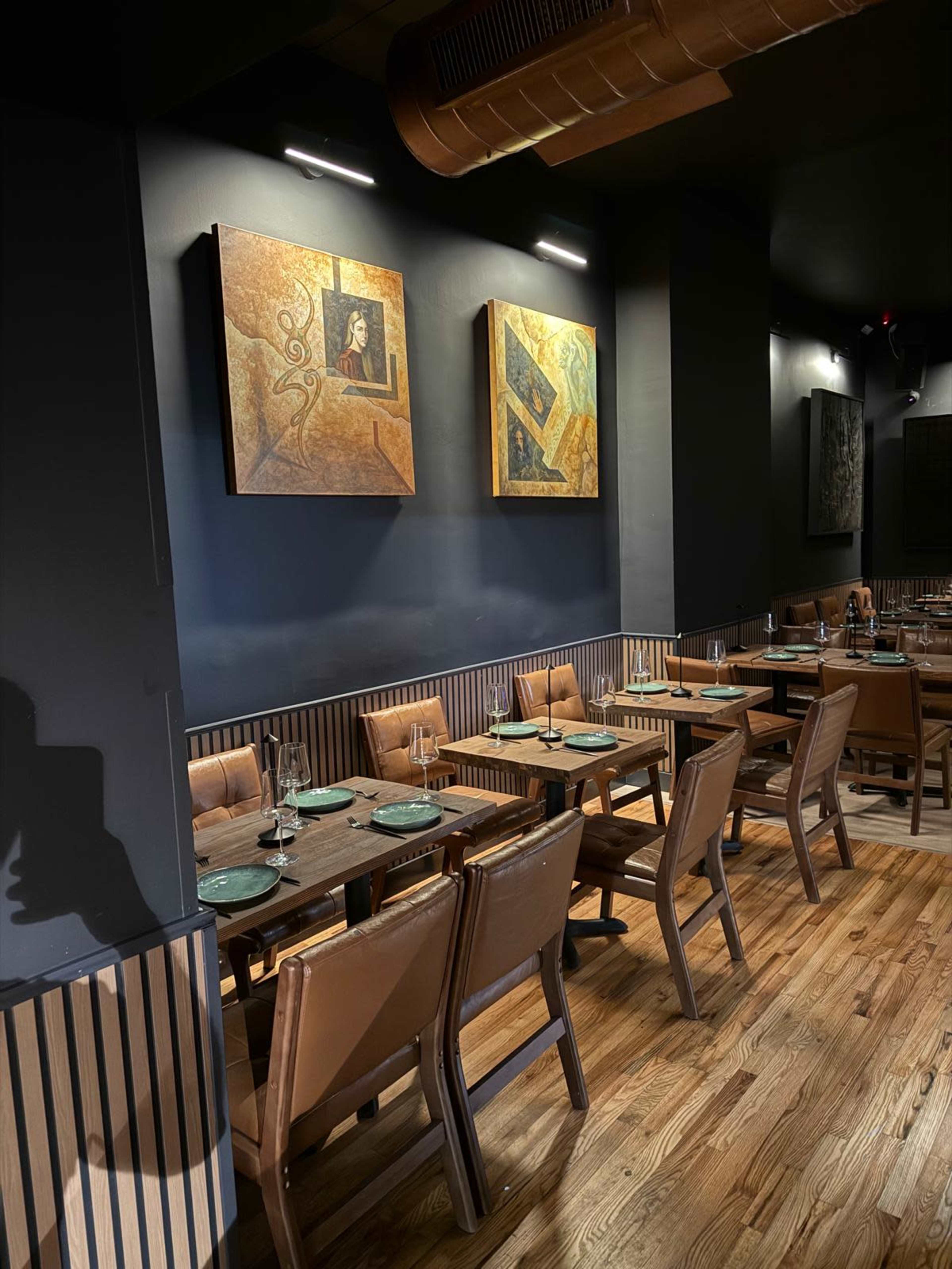 The image shows a modern restaurant interior featuring wooden tables and leather chairs, with abstract art on the dark walls.