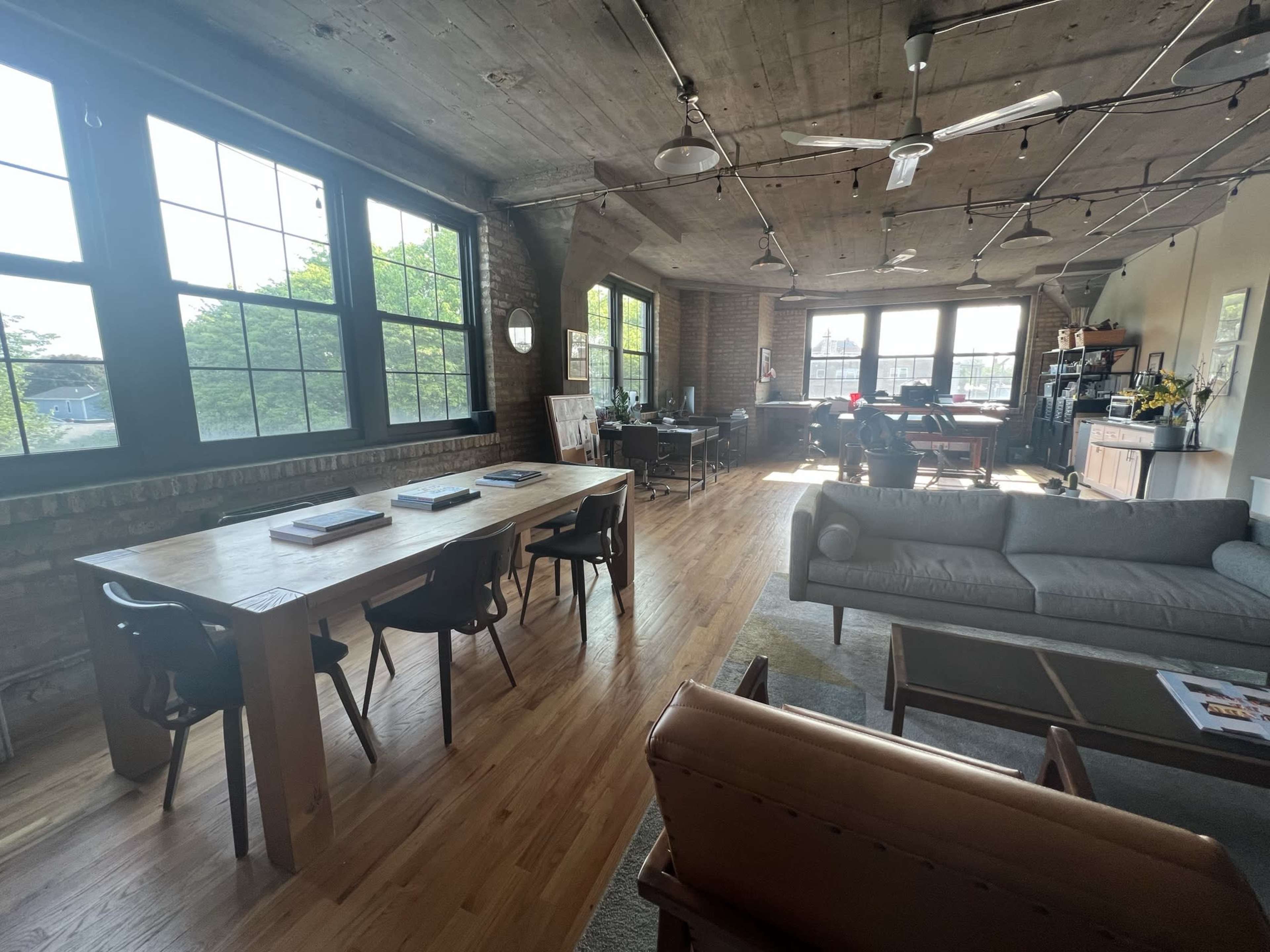 An open-concept loft space featuring a wooden dining table, a sofa, and a workspace with large windows allowing natural light to fill the room.