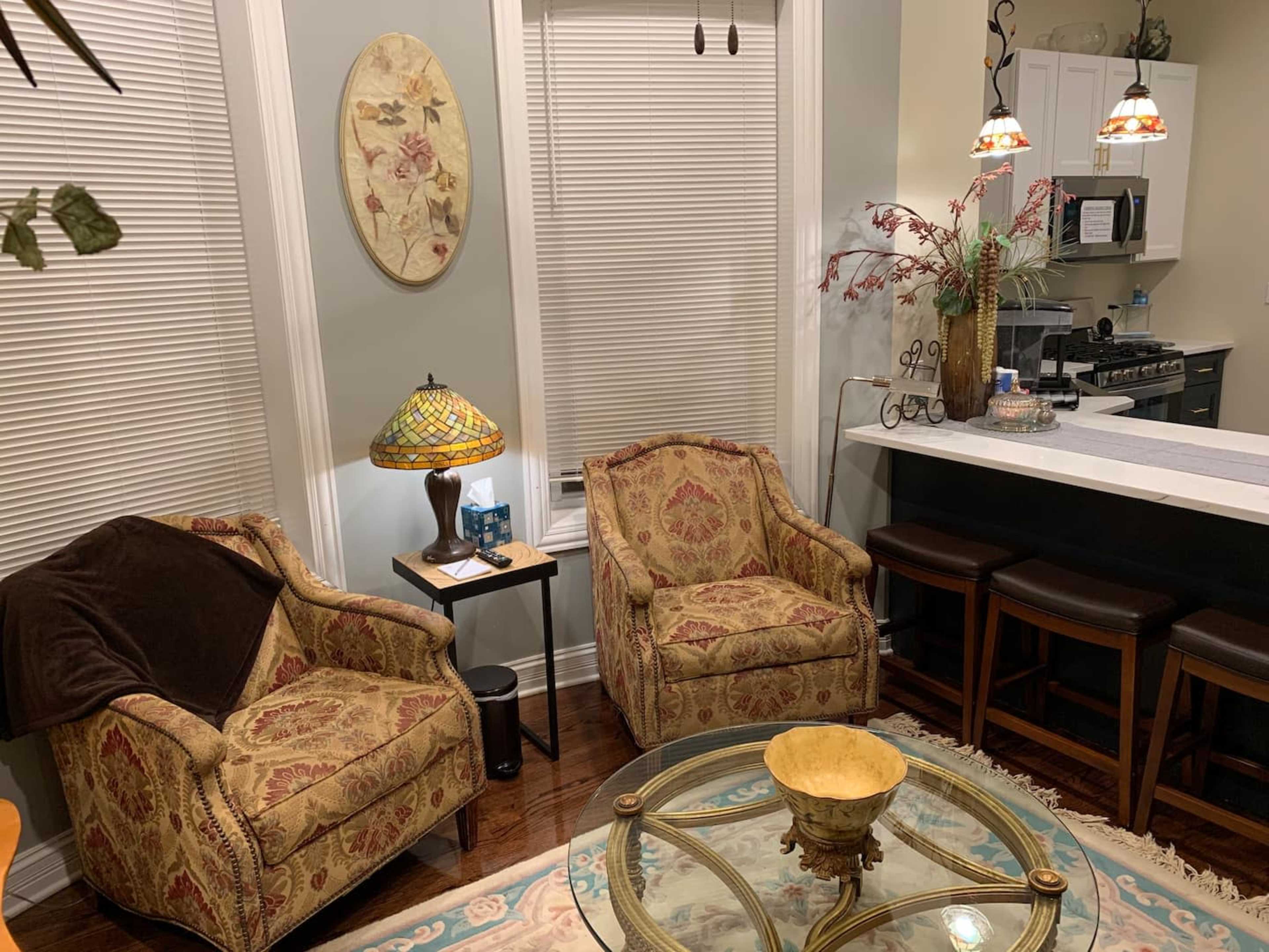 The image shows a cozy living space with two patterned armchairs, a glass coffee table, and a kitchen area in the background with bar seating.