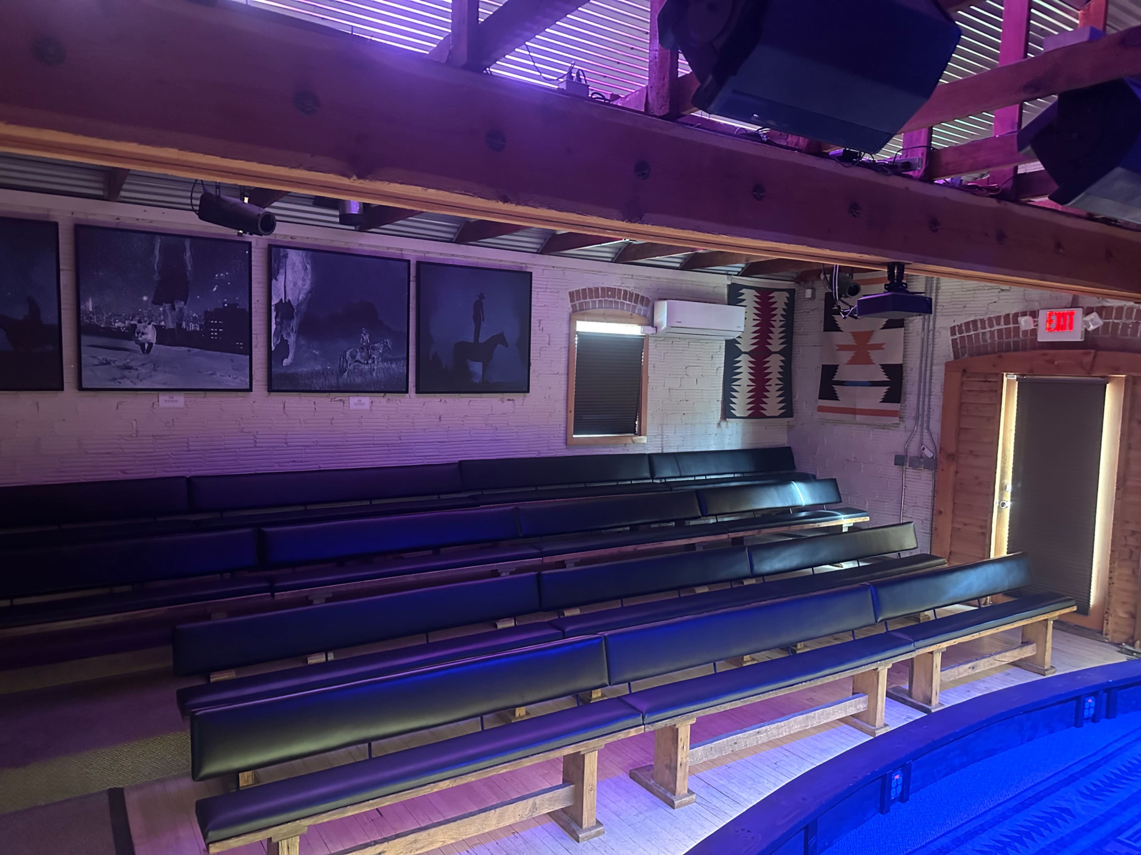 A sparsely furnished performance space with black benches arranged in rows, surrounded by wooden walls and framed black-and-white photographs.