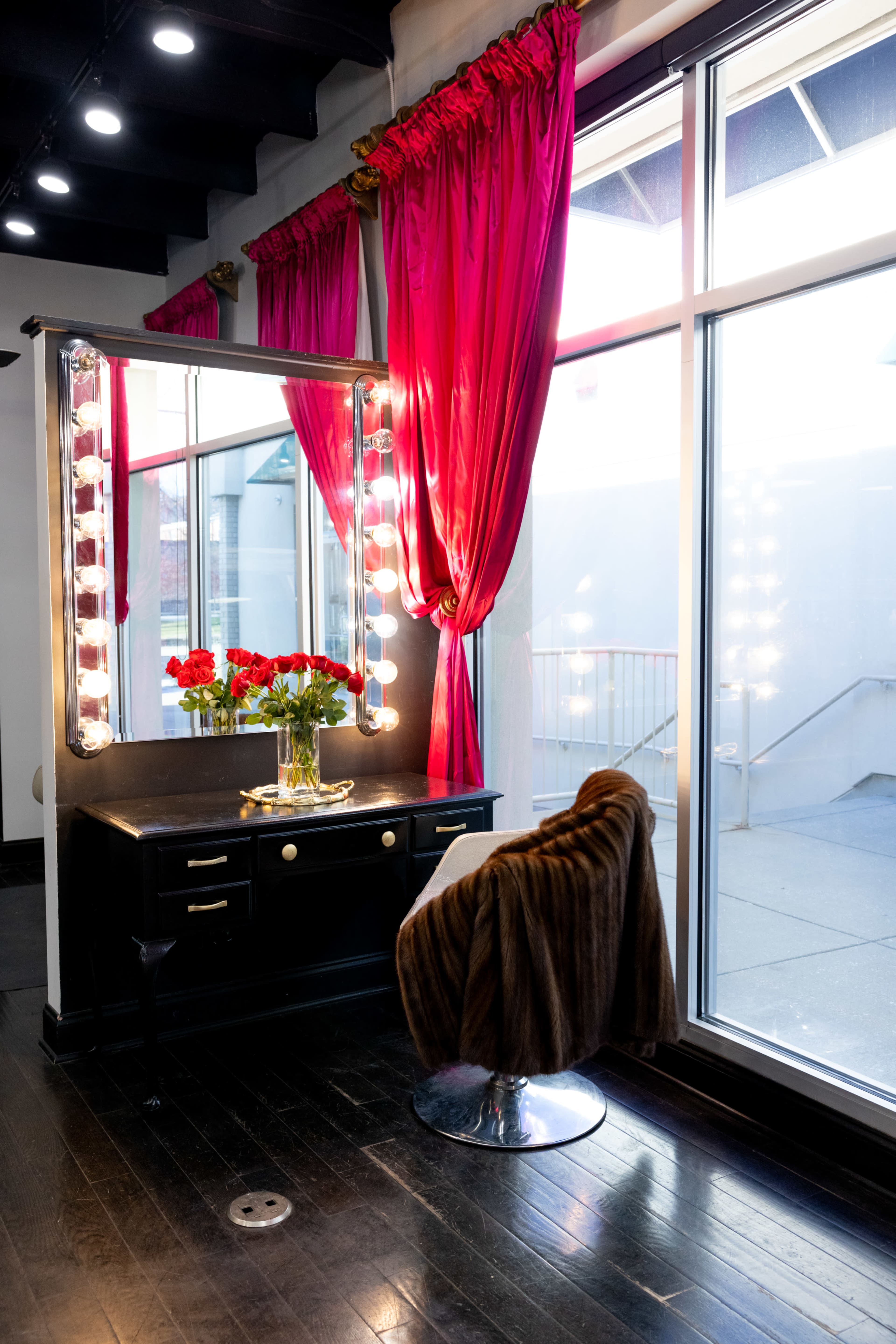The image features a makeup vanity with bright lights, a bouquet of red roses, and a plush brown chair, all set against red curtains and large windows.