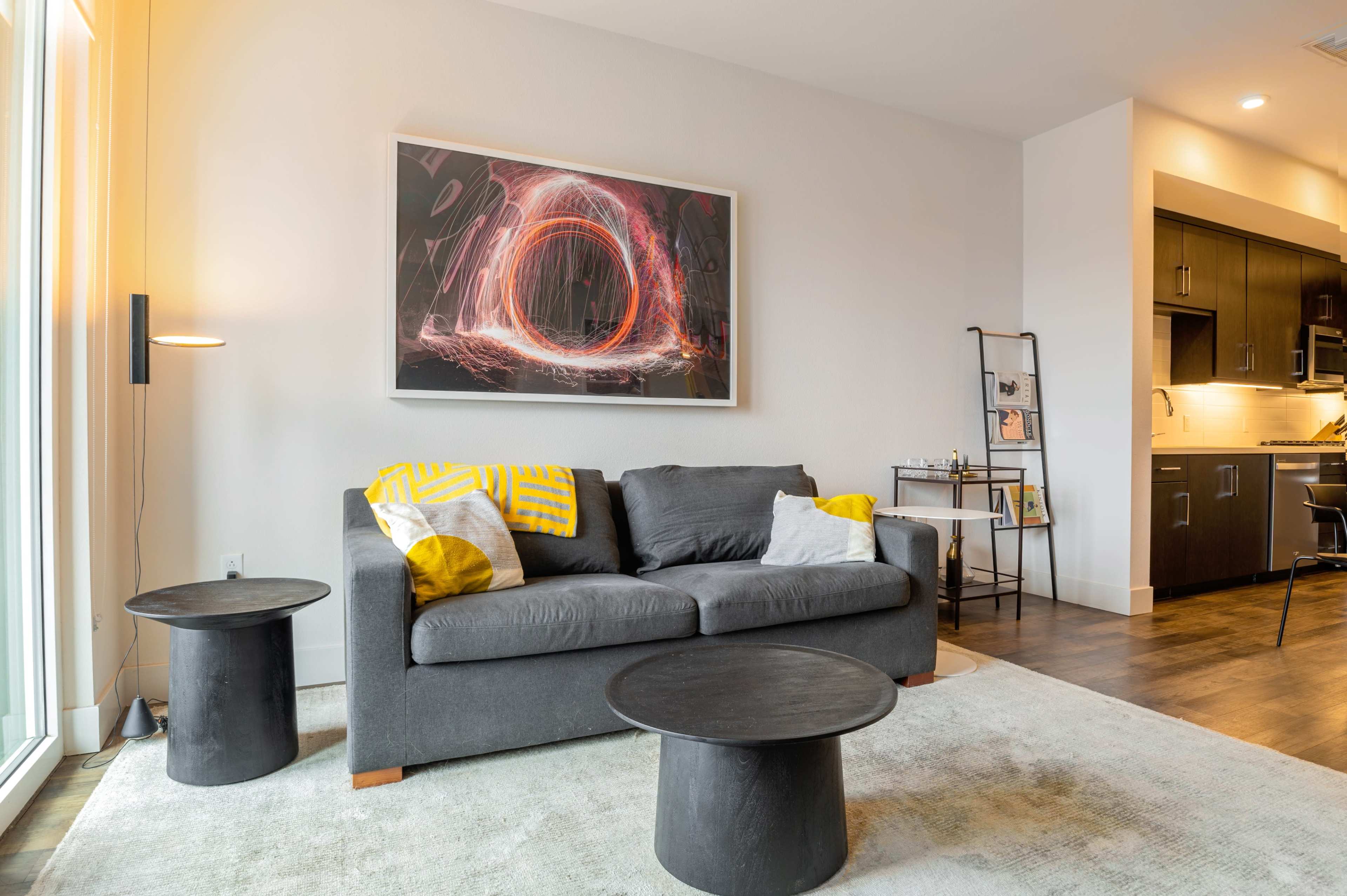 A modern living room features a gray sofa with yellow and white pillows, two black circular coffee tables, and a large framed artwork on the wall, alongside a kitchenette.