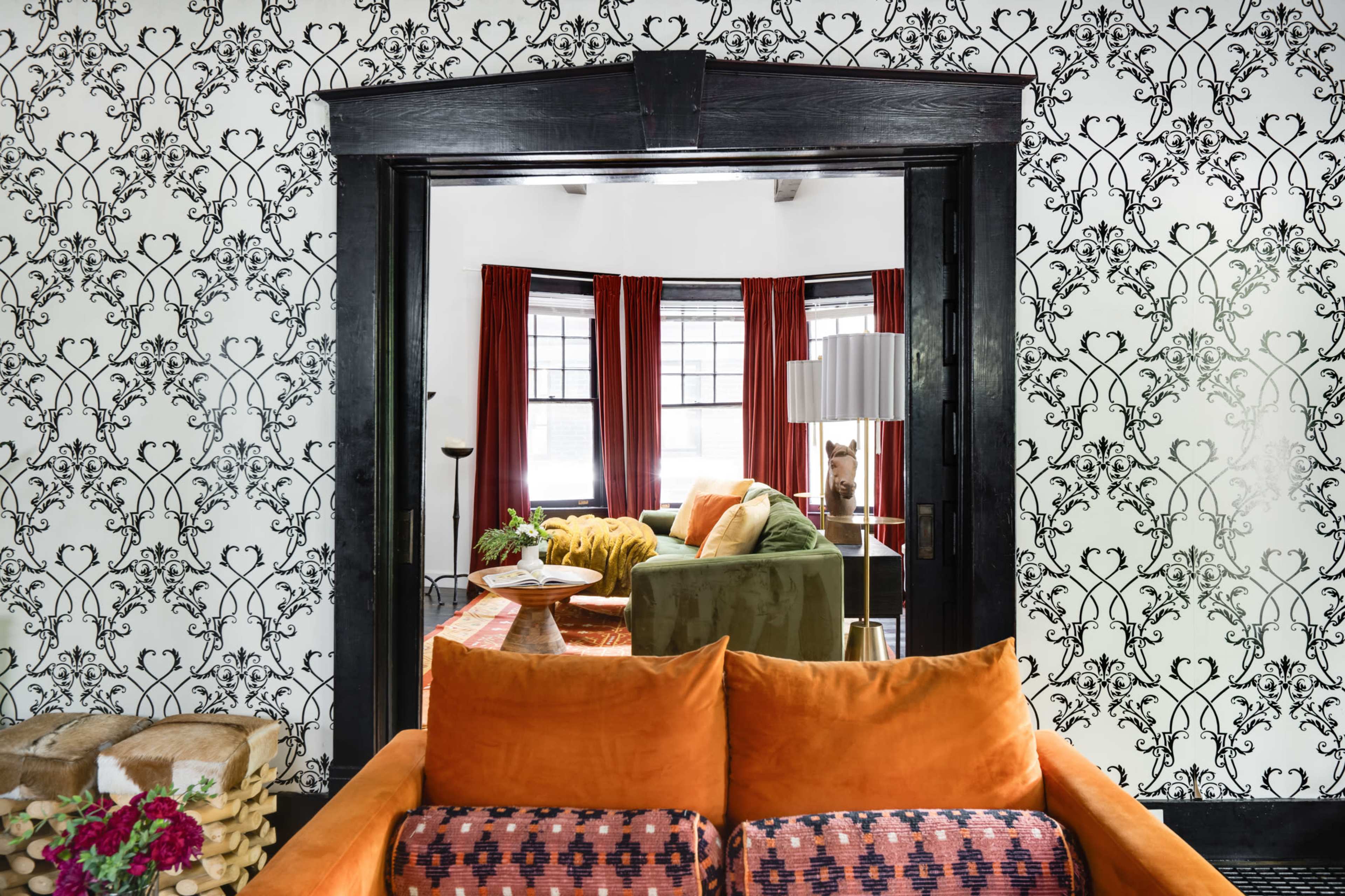 The image shows a living room with a patterned black-and-white wallpaper, orange and green furniture, large windows with red curtains, and a stylish area rug.