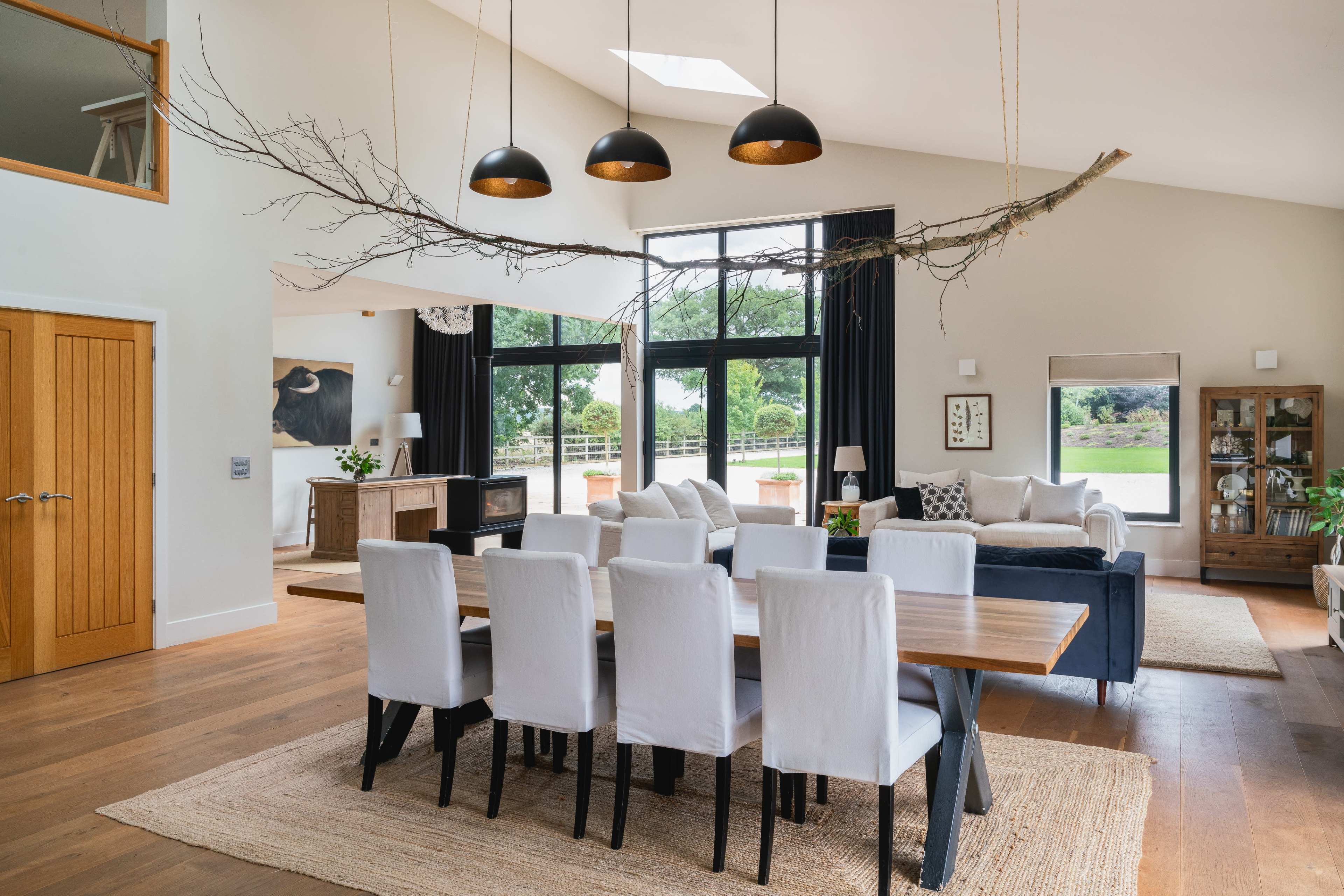 A spacious, modern dining area features a long wooden table surrounded by white chairs, with a branch light fixture overhead and large windows allowing natural light to fill the room.
