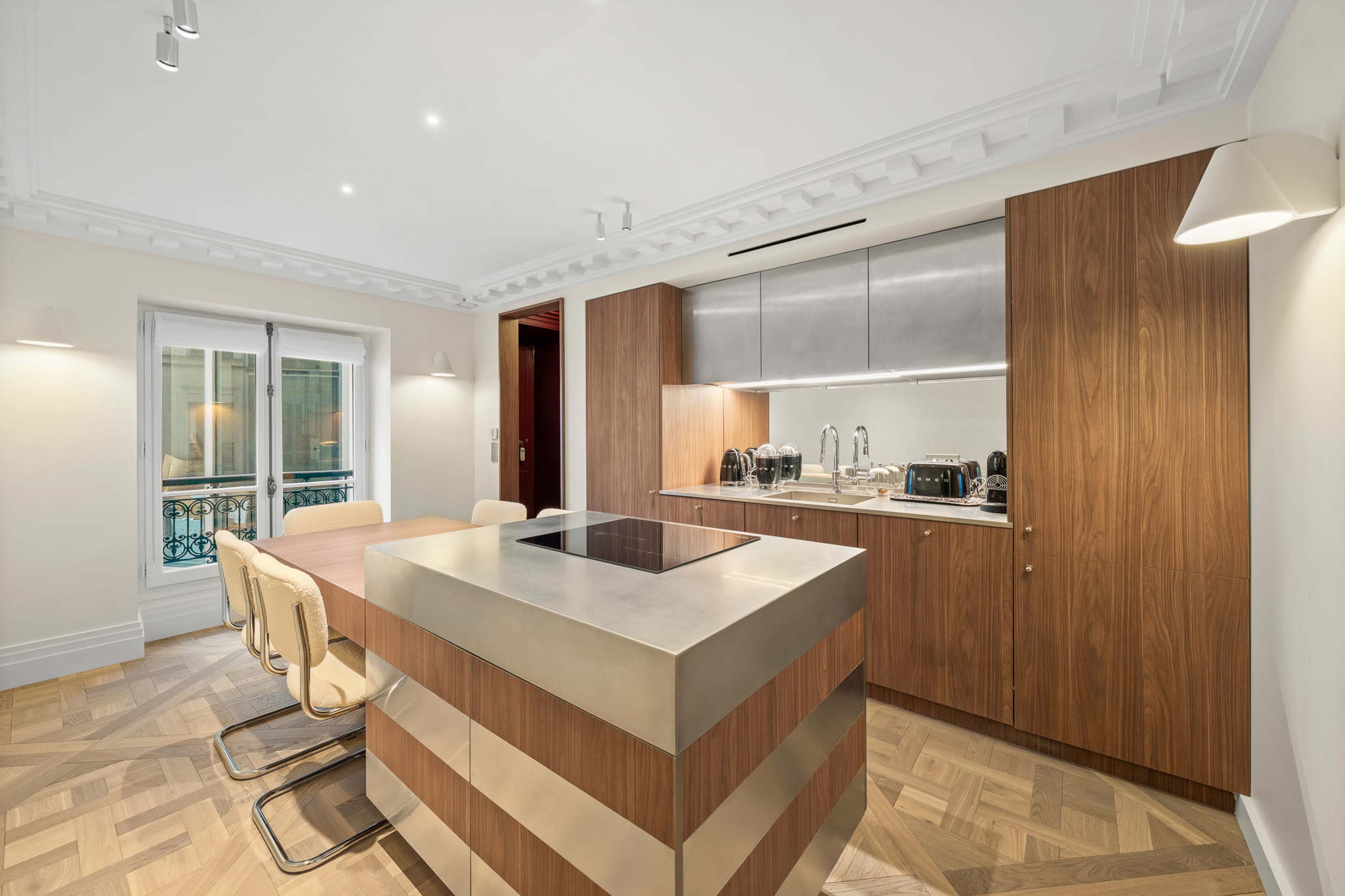 The image shows a modern kitchen with a sleek island, wooden cabinetry, and a dining area featuring beige chairs.
