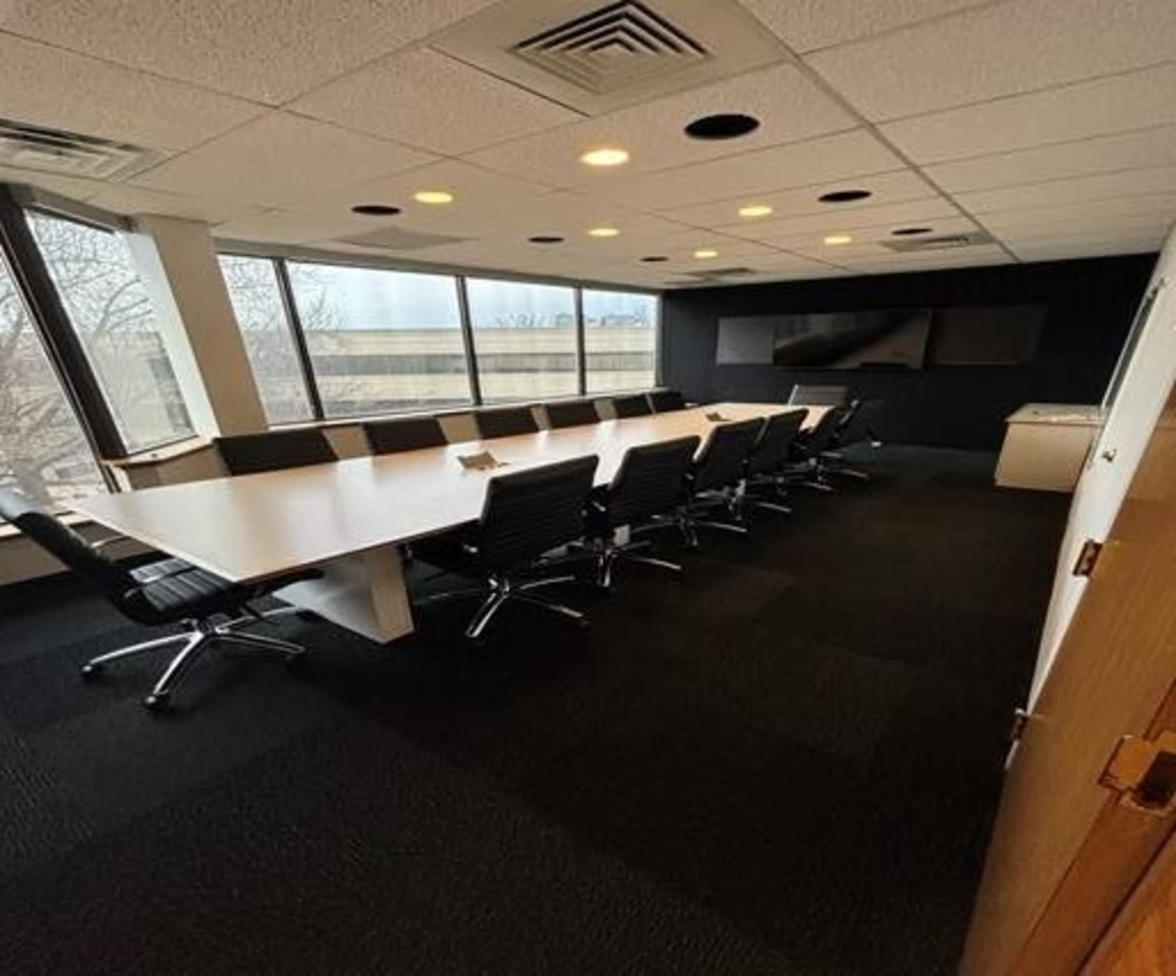 A large conference room is furnished with a long table and several black ergonomic chairs, featuring large windows and modern lighting.
