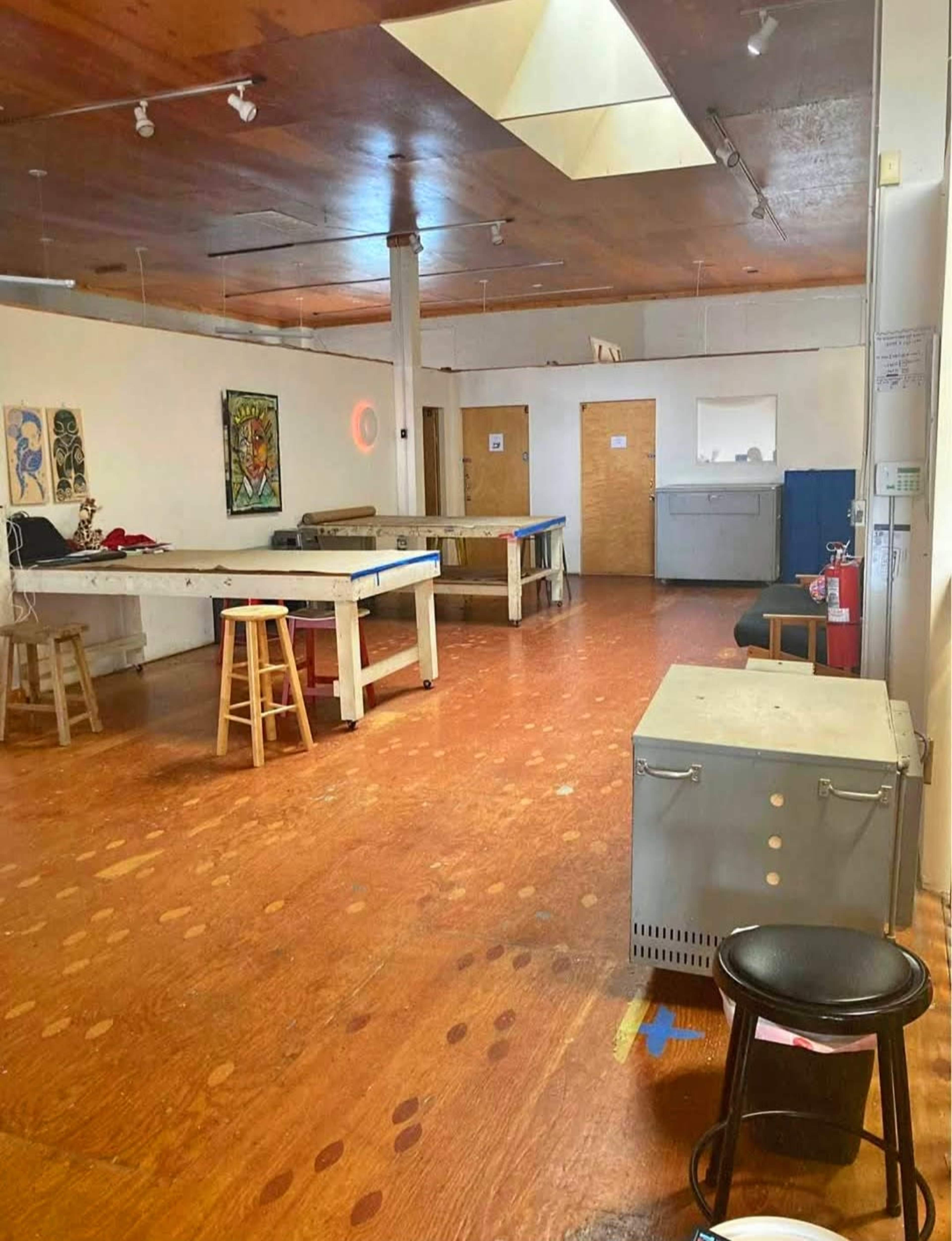 The image depicts a spacious, empty room with wooden flooring, two tables, and various pieces of furniture, including stools and a storage container.