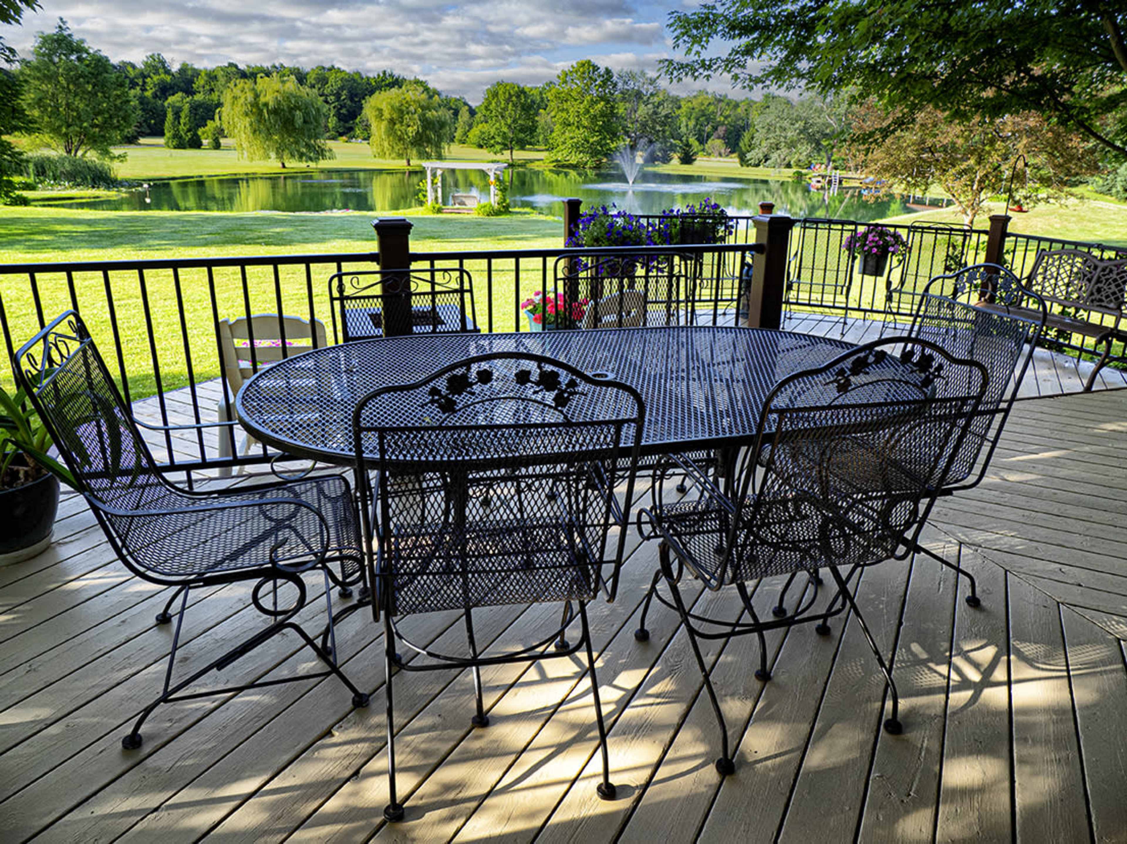 A metal patio set with six chairs surrounds a round table on a deck overlooking a tranquil pond and lush greenery.
