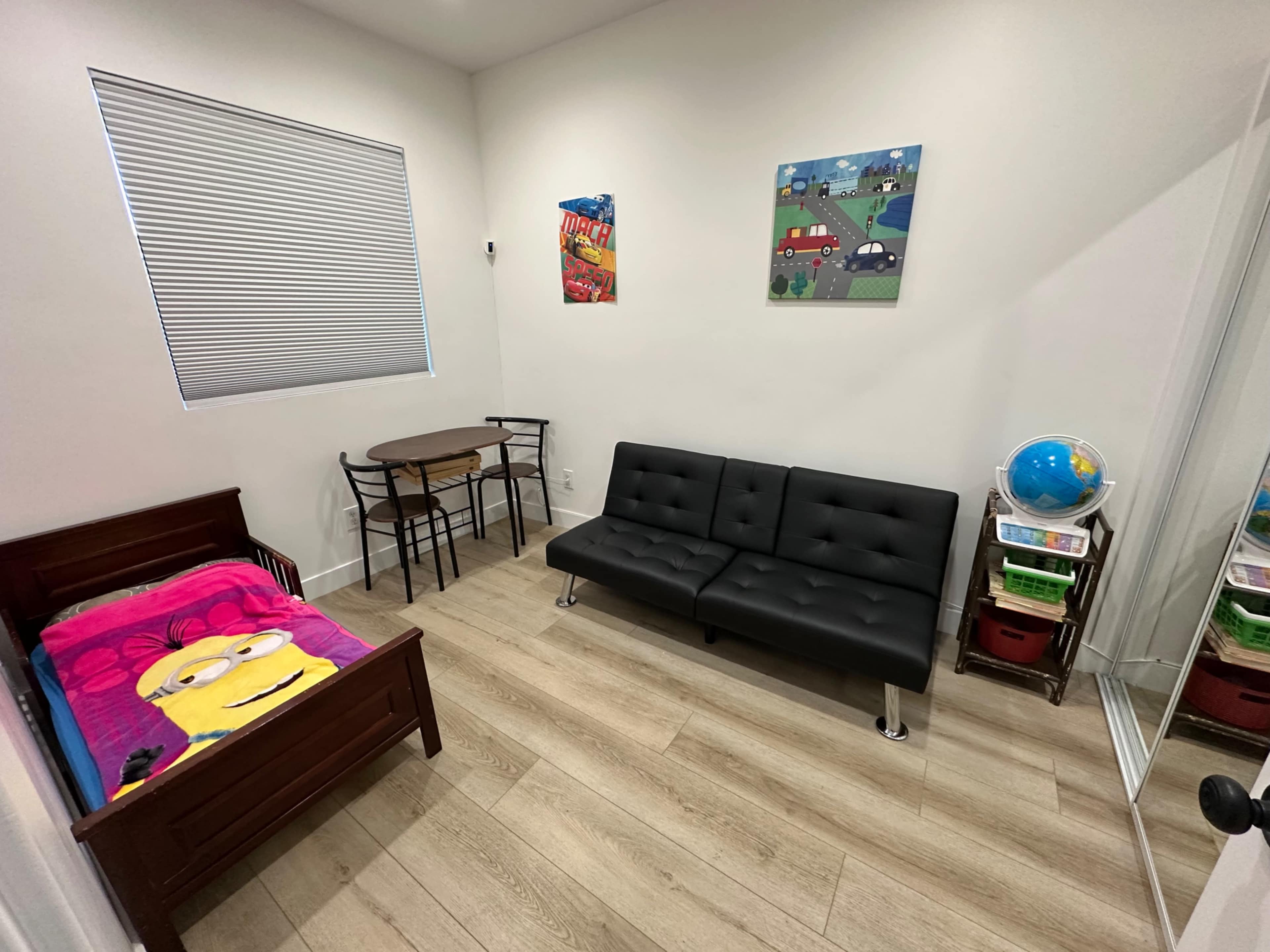 The image shows a small, well-lit room featuring a bed with a colorful blanket, a black sofa, a table with chairs, and a globe on a shelf beside a mirror.