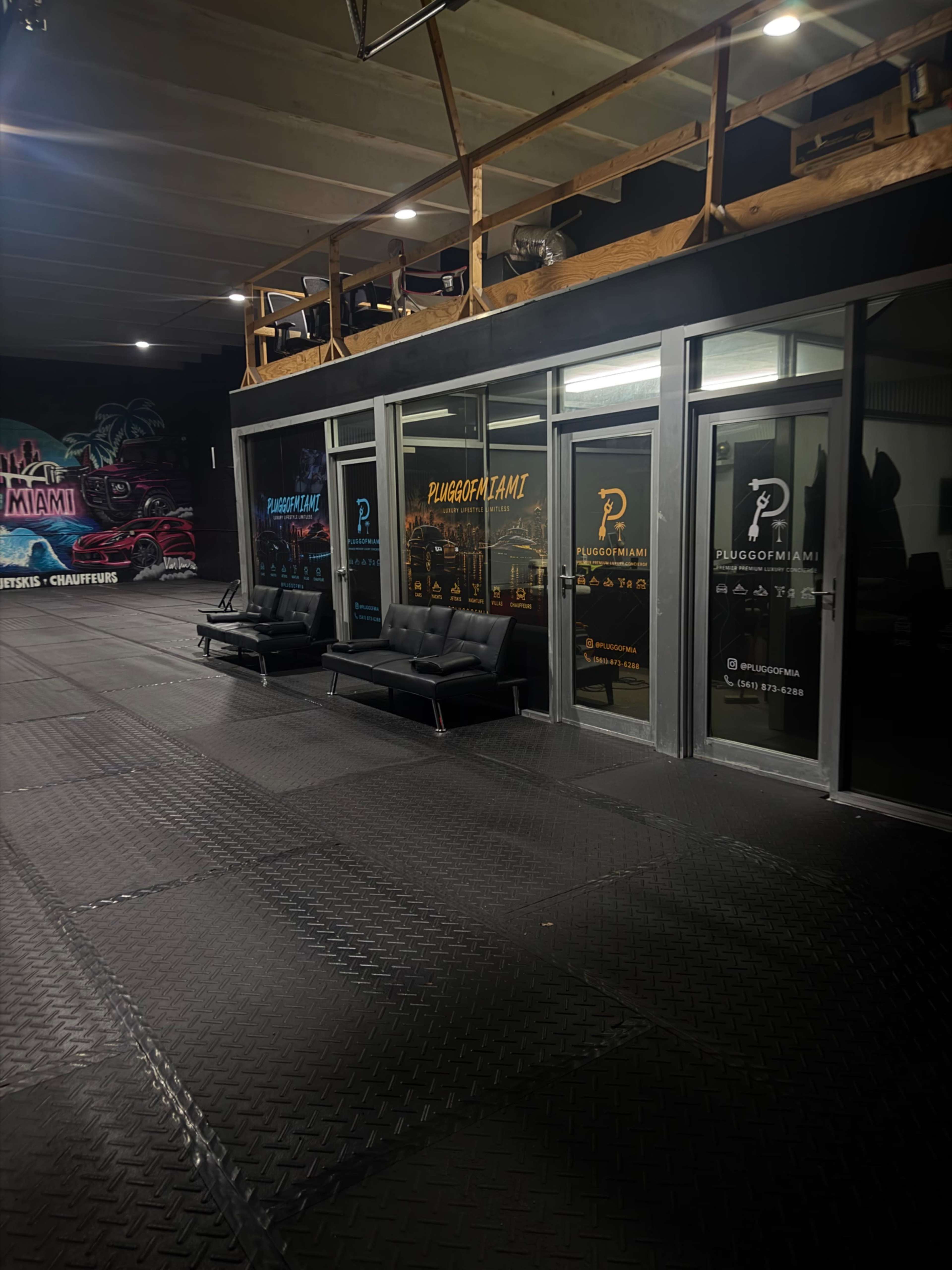 The image shows a gym or fitness studio interior with large glass windows and black rubber flooring, featuring two black sofas outside the entrance.