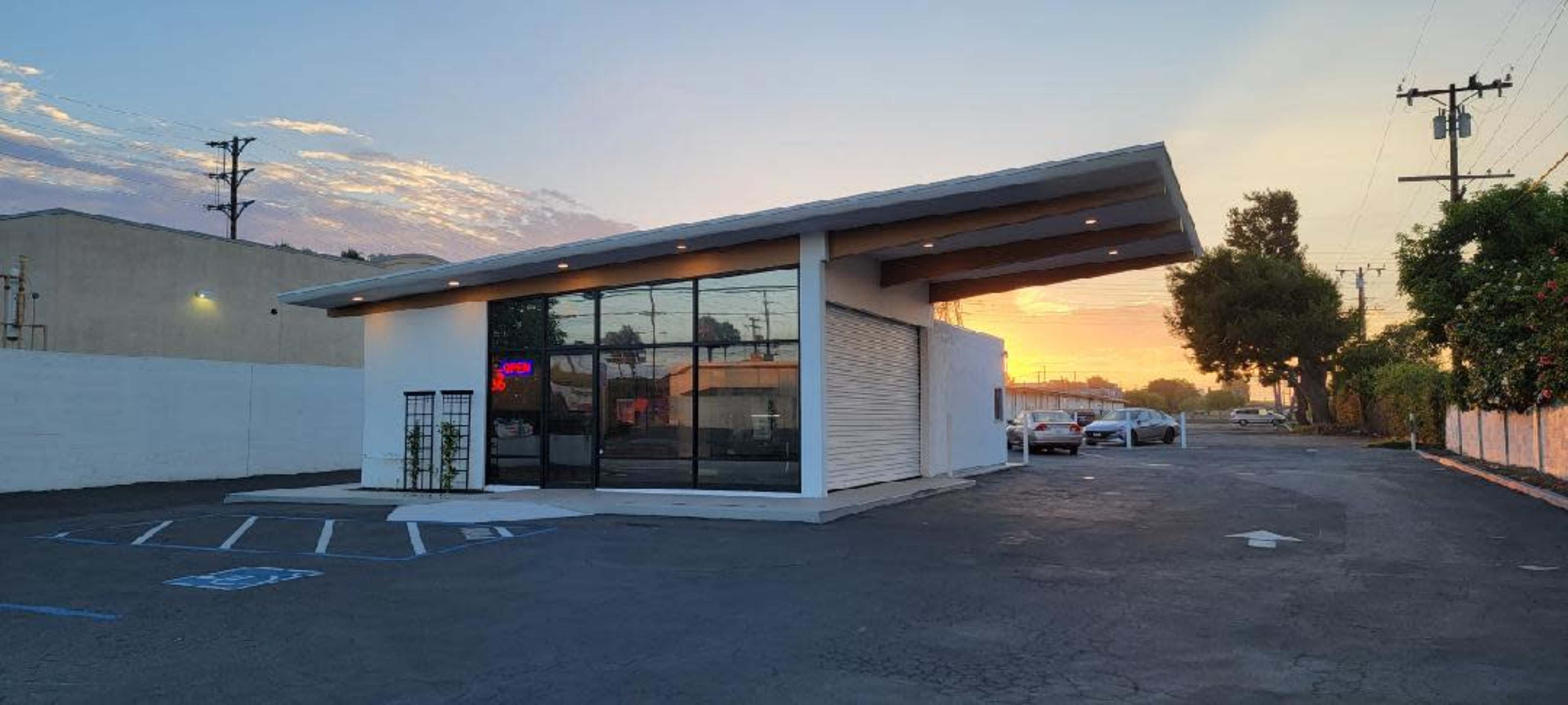 A modern, single-story building with large glass windows and a flat roof is situated in a parking lot at sunset.